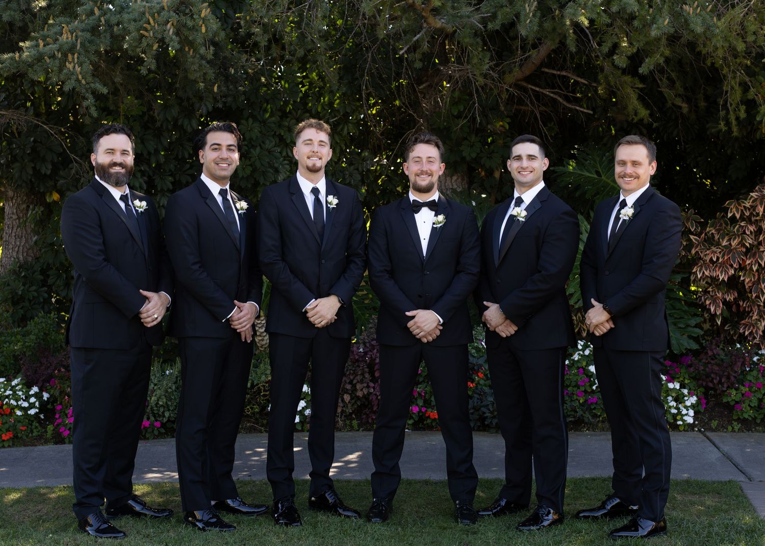 Groomsmen in black tuxedos line up for formal wedding photos outdoors.