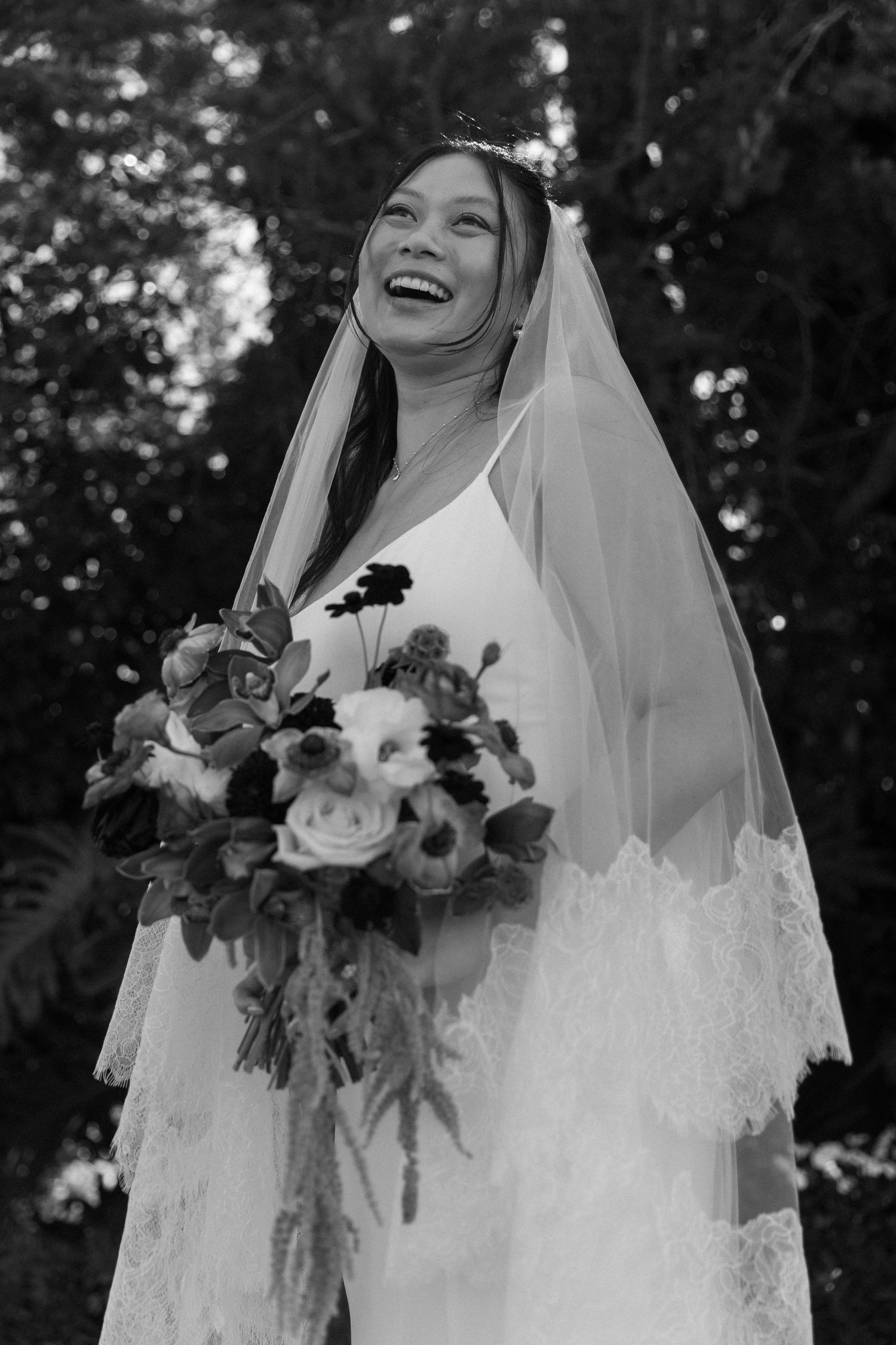 Bride in lace veil holding a beautiful bouquet of mixed flowers outdoors.