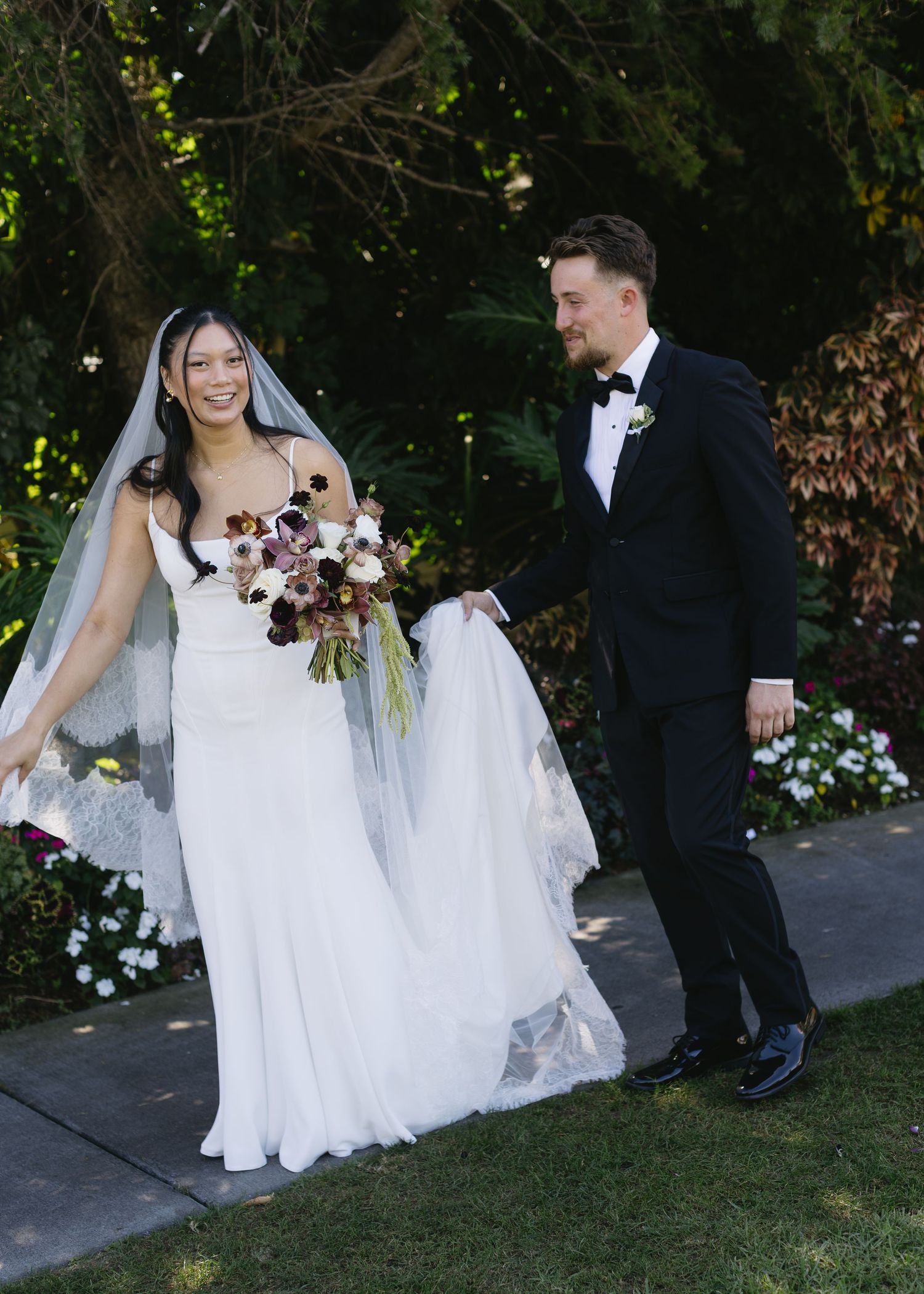 A couple in wedding attire walks together on a garden path with flowing white dress and veil.