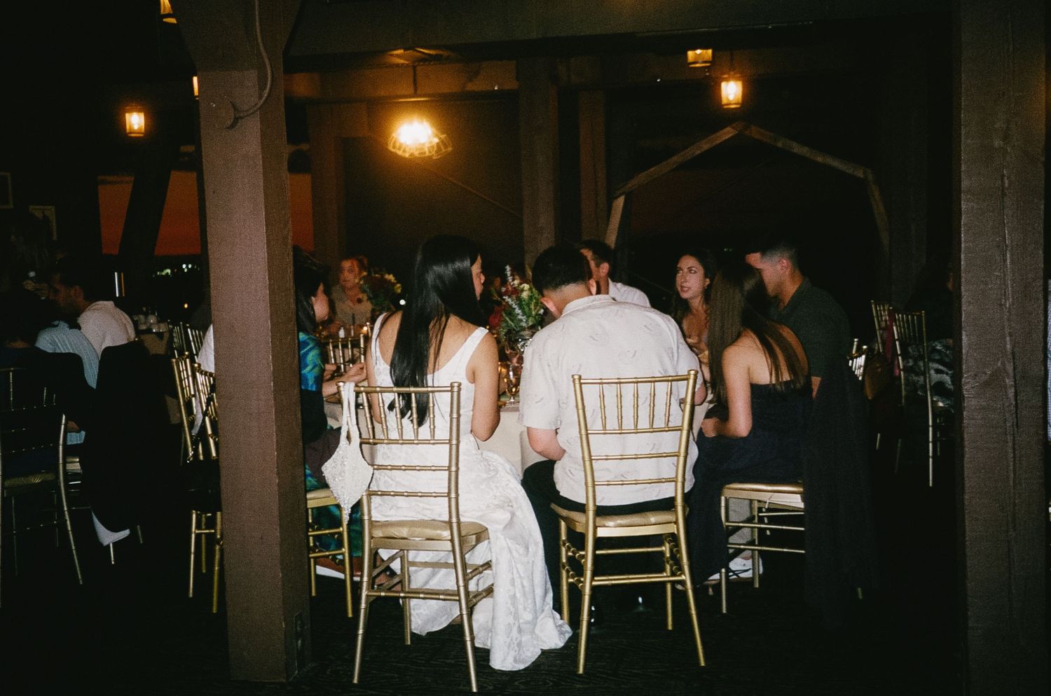Wedding guests seated at round tables in a dimly lit reception venue with warm glowing lights overhead.