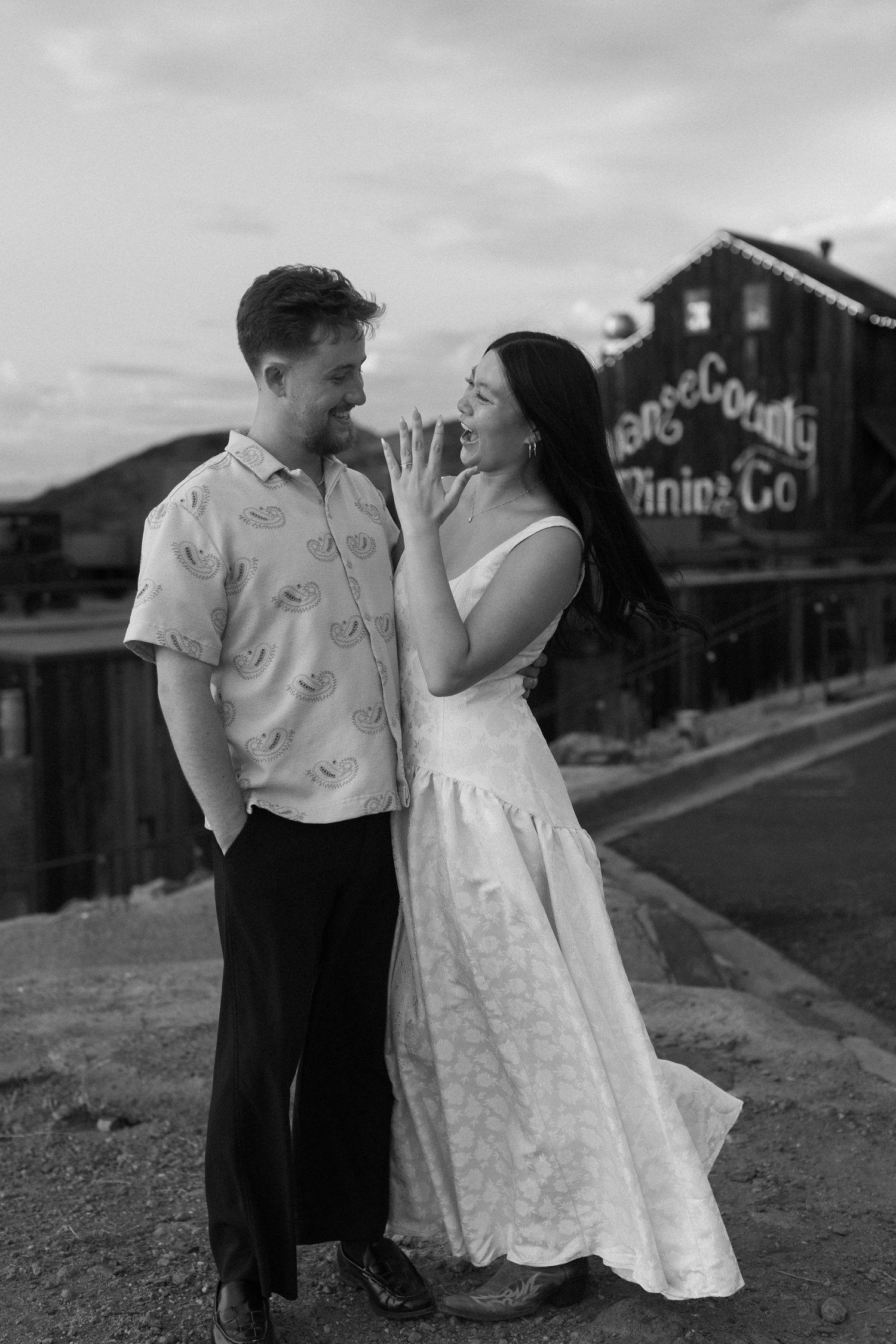 Romantic couple poses for photos near rustic buildings at sunset.