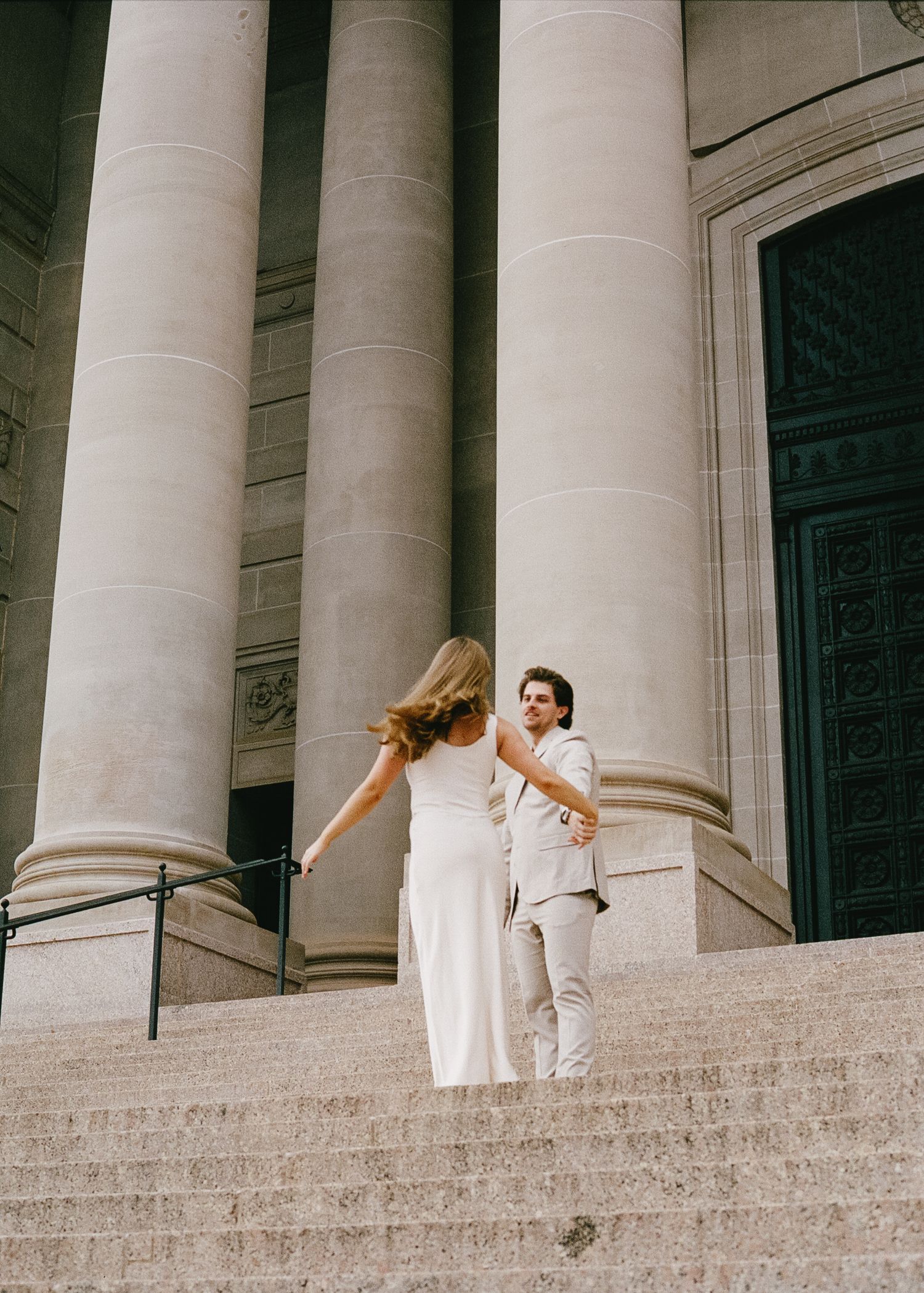 A couple in white and beige attire posing on grand stone steps with classical columns.