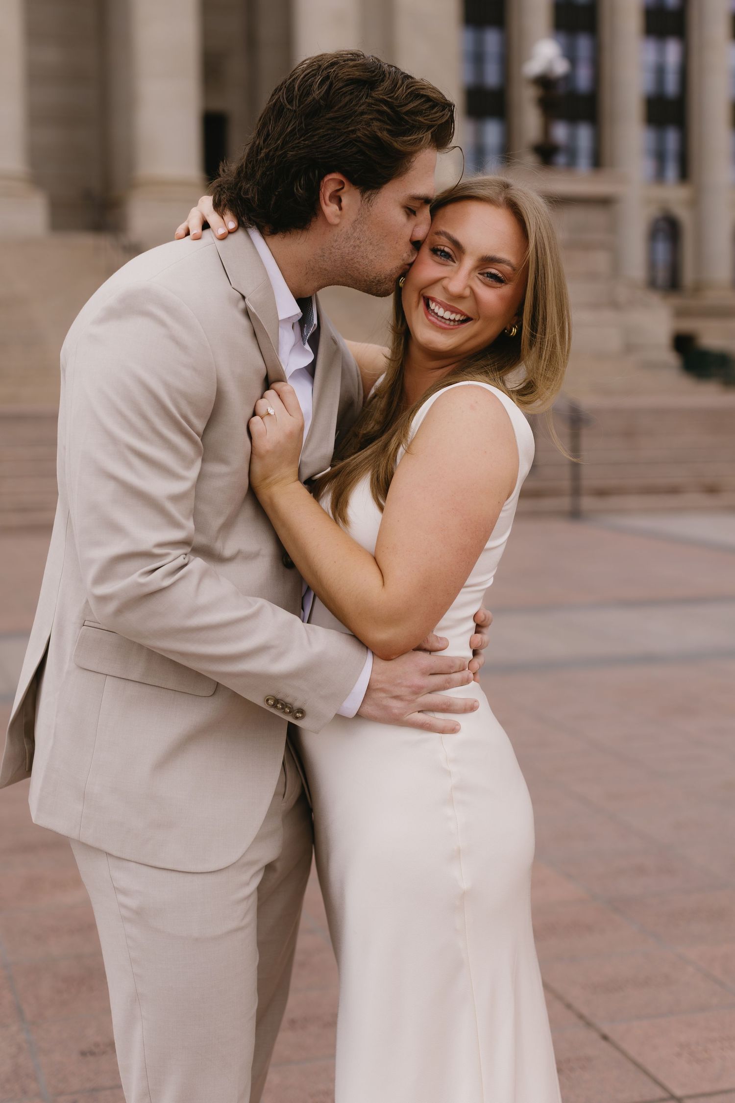A couple in beige and white attire shares tender moments outside a classical building.