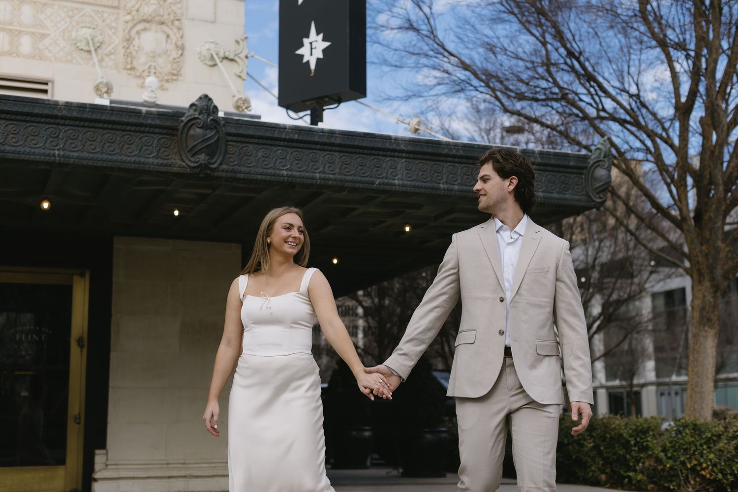 A couple in light-colored formal wear hold hands while walking outside a building.