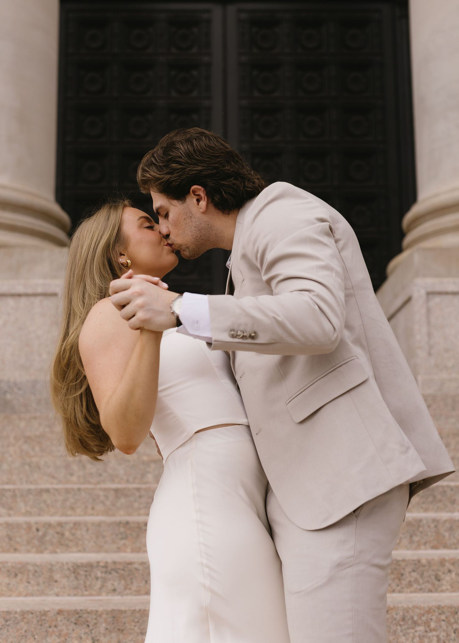 A couple in light formal wear shares a romantic kiss on stone steps.