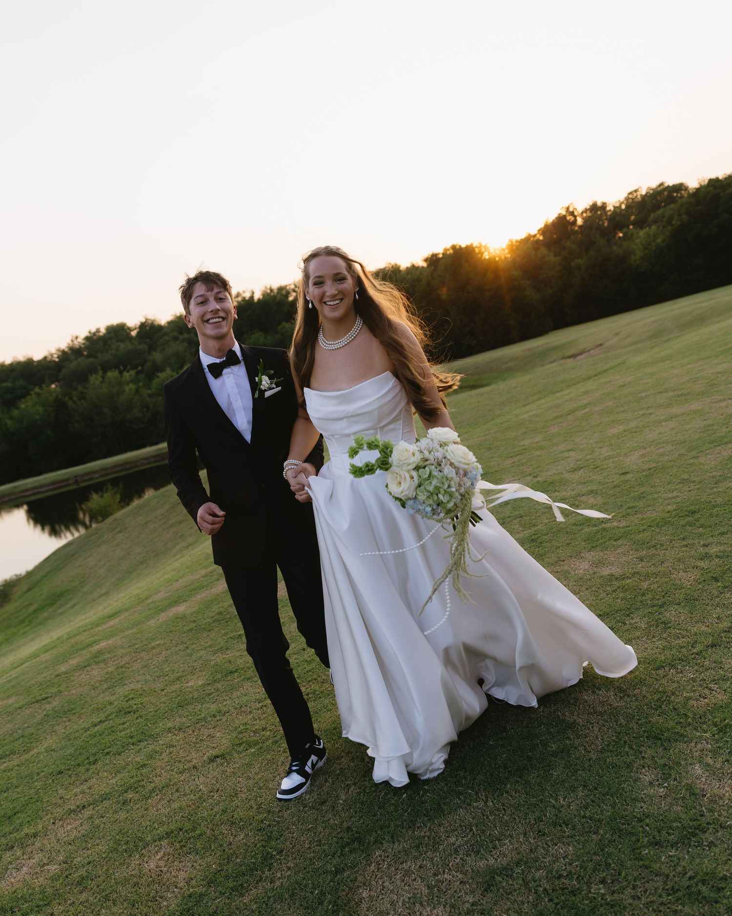 Newlyweds walking playfully across a grassy field at sunset.
