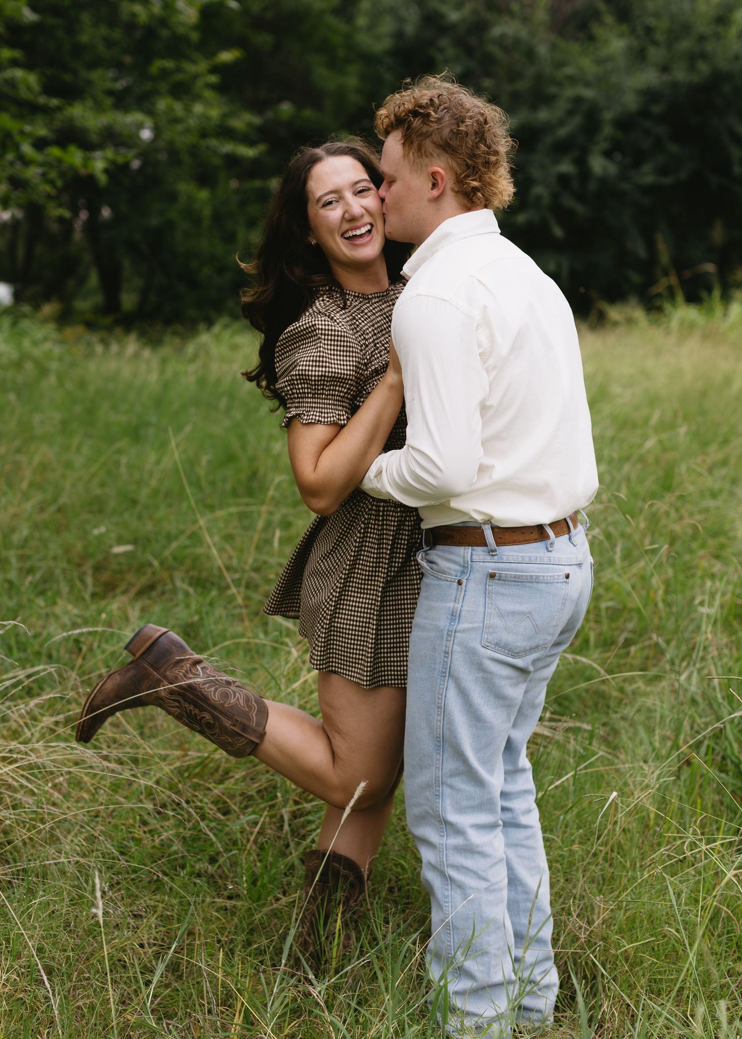 Couple embraces playfully in a grassy meadow wearing country-western inspired outfits.