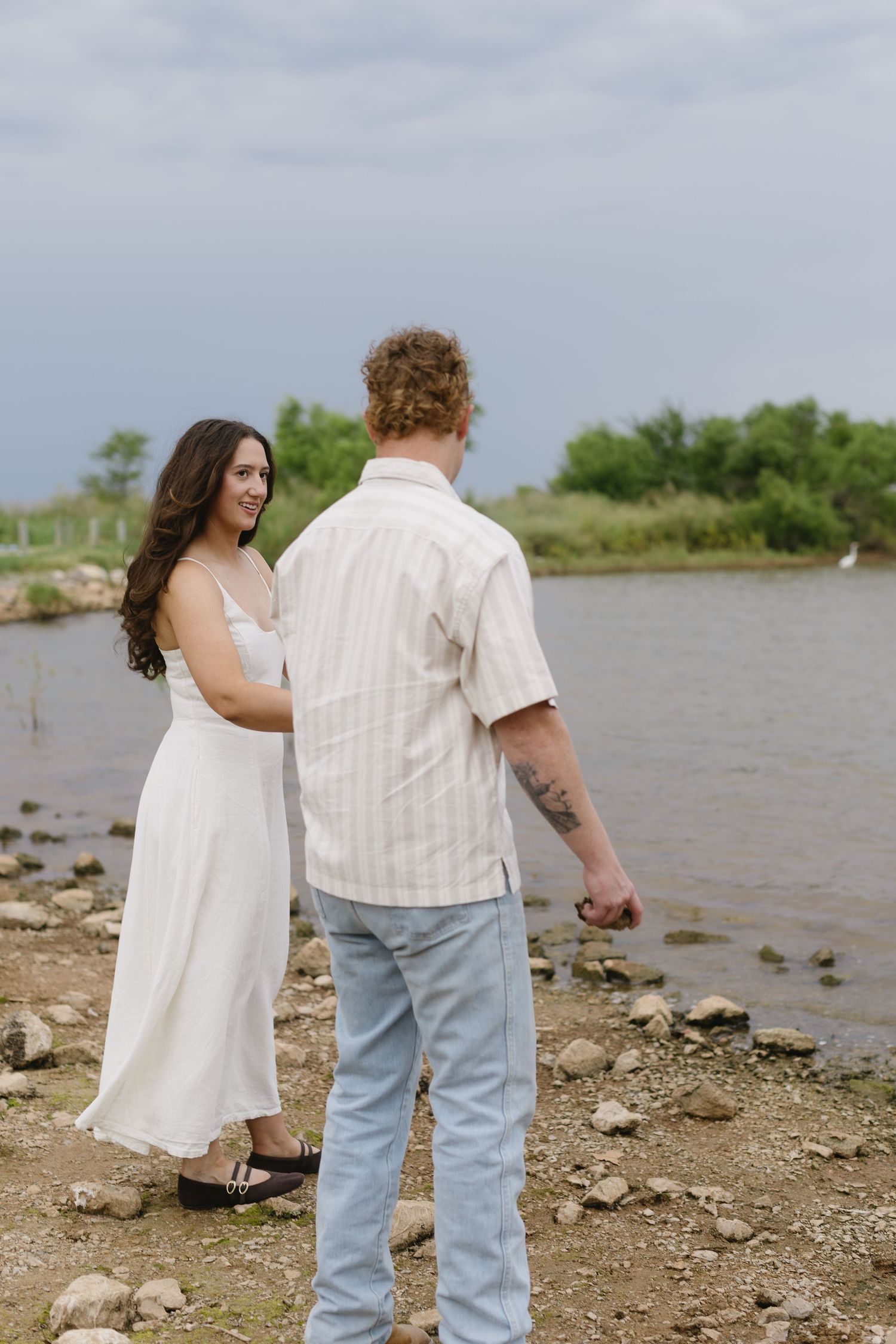 A couple in coordinating white outfits walks along a rocky shoreline on an overcast day.
