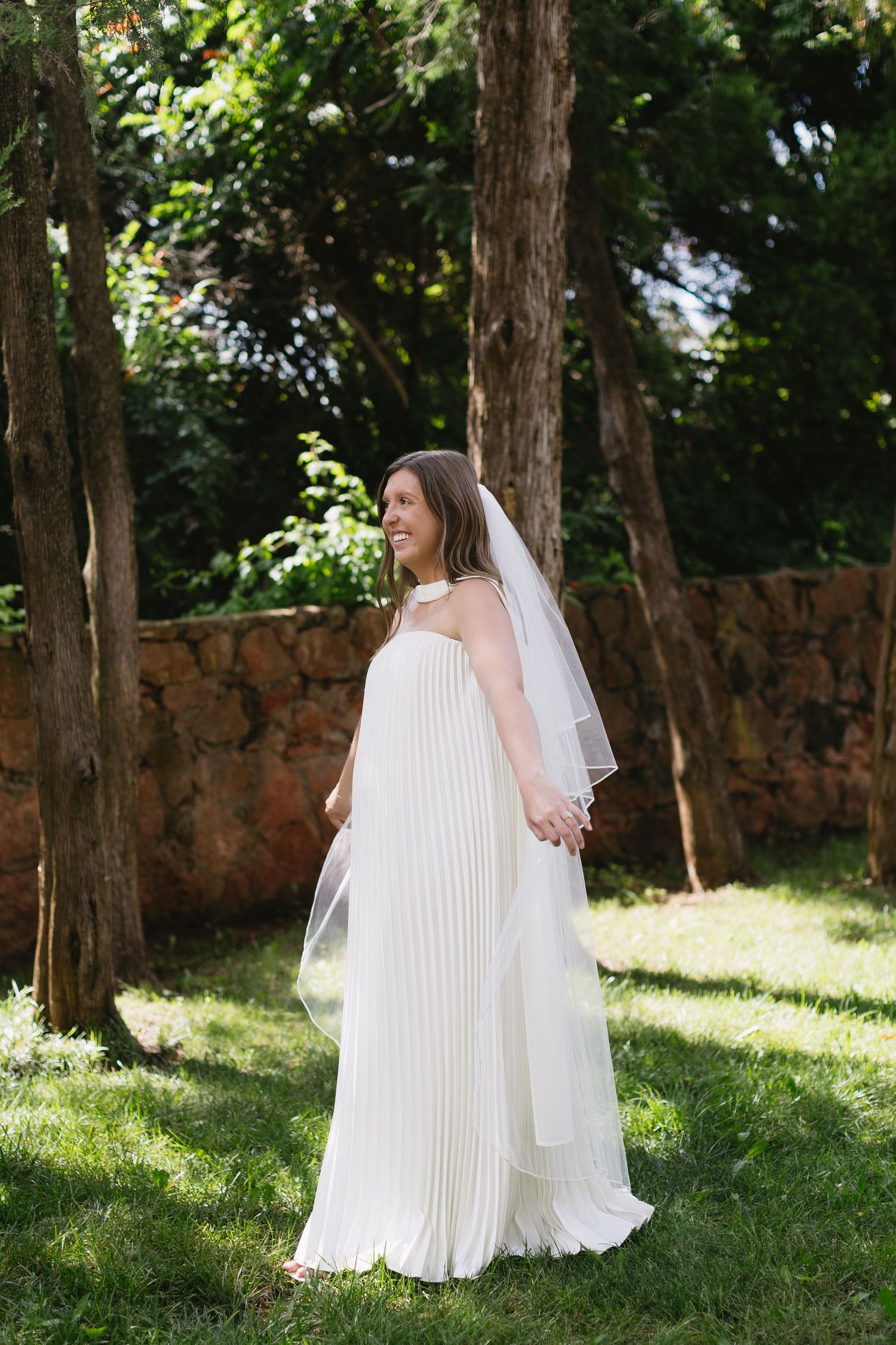 Ethereal white pleated wedding dress photographed in a sunlit garden setting.