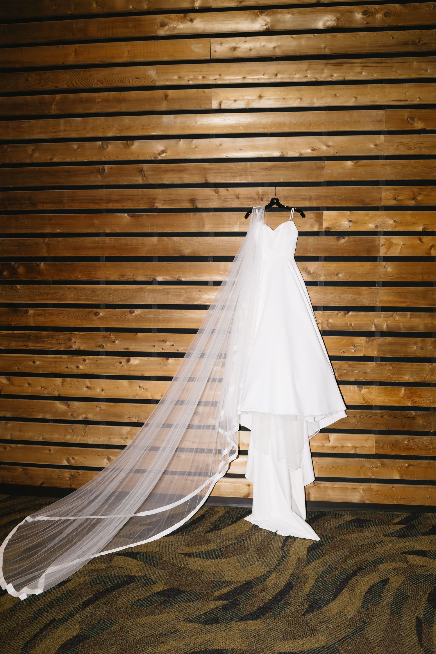 Elegant white wedding dress and flowing veil laid out against rustic wooden slat wall backdrop.