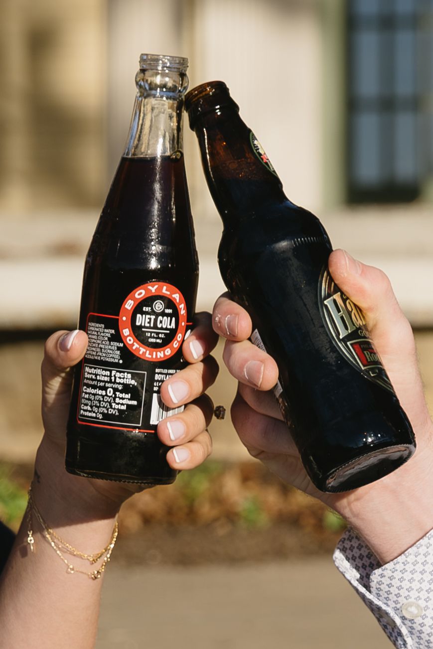 Two hands clink glass bottles of dark soda or beer together in a celebratory toast against a blurred background.