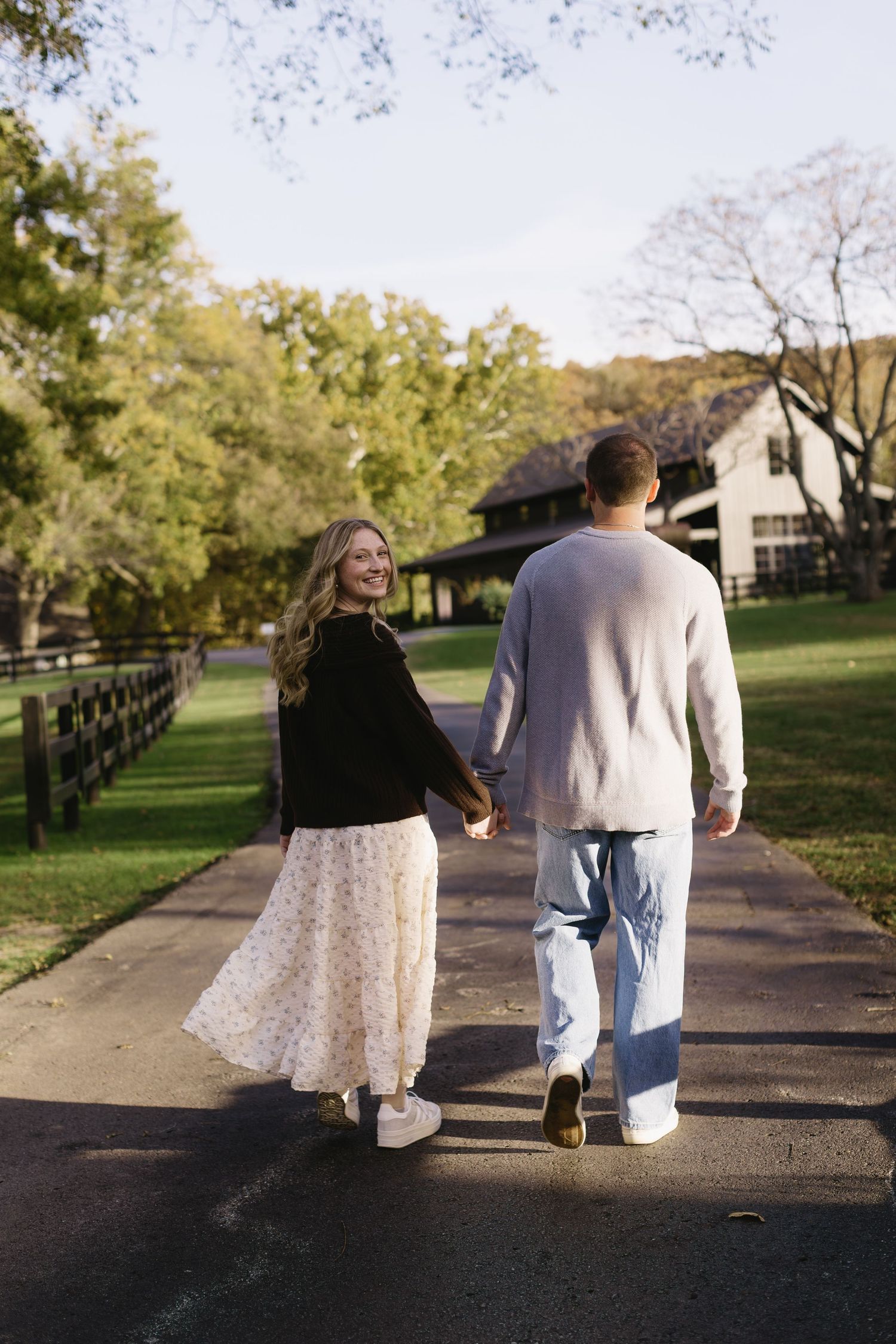 A couple walking hand in hand on a path with white fencing and autumn trees in the background.