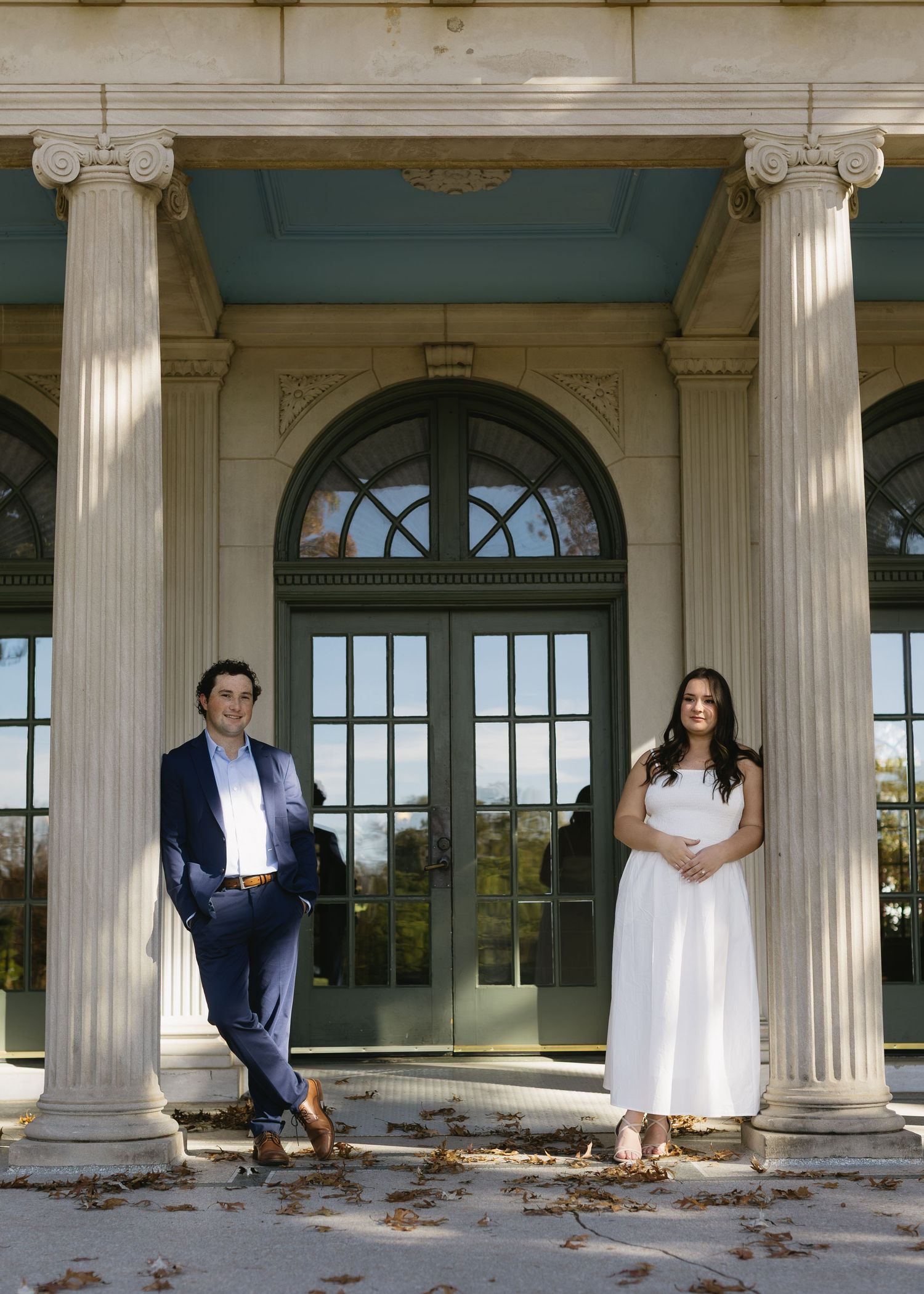 A couple poses playfully between elegant white columns of a classical building.