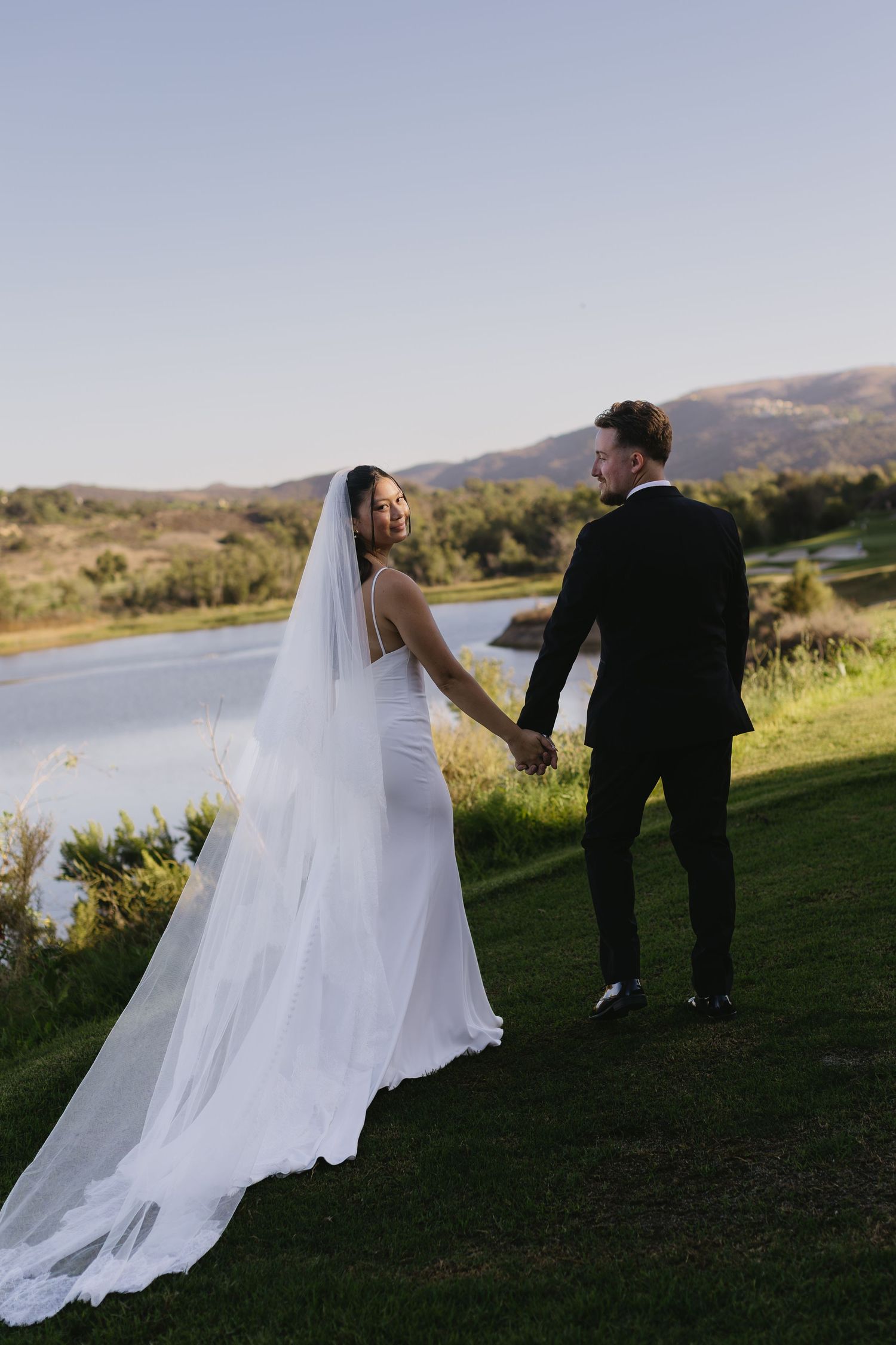 A wedding couple walks along a grassy hillside overlooking a scenic lake during golden hour.