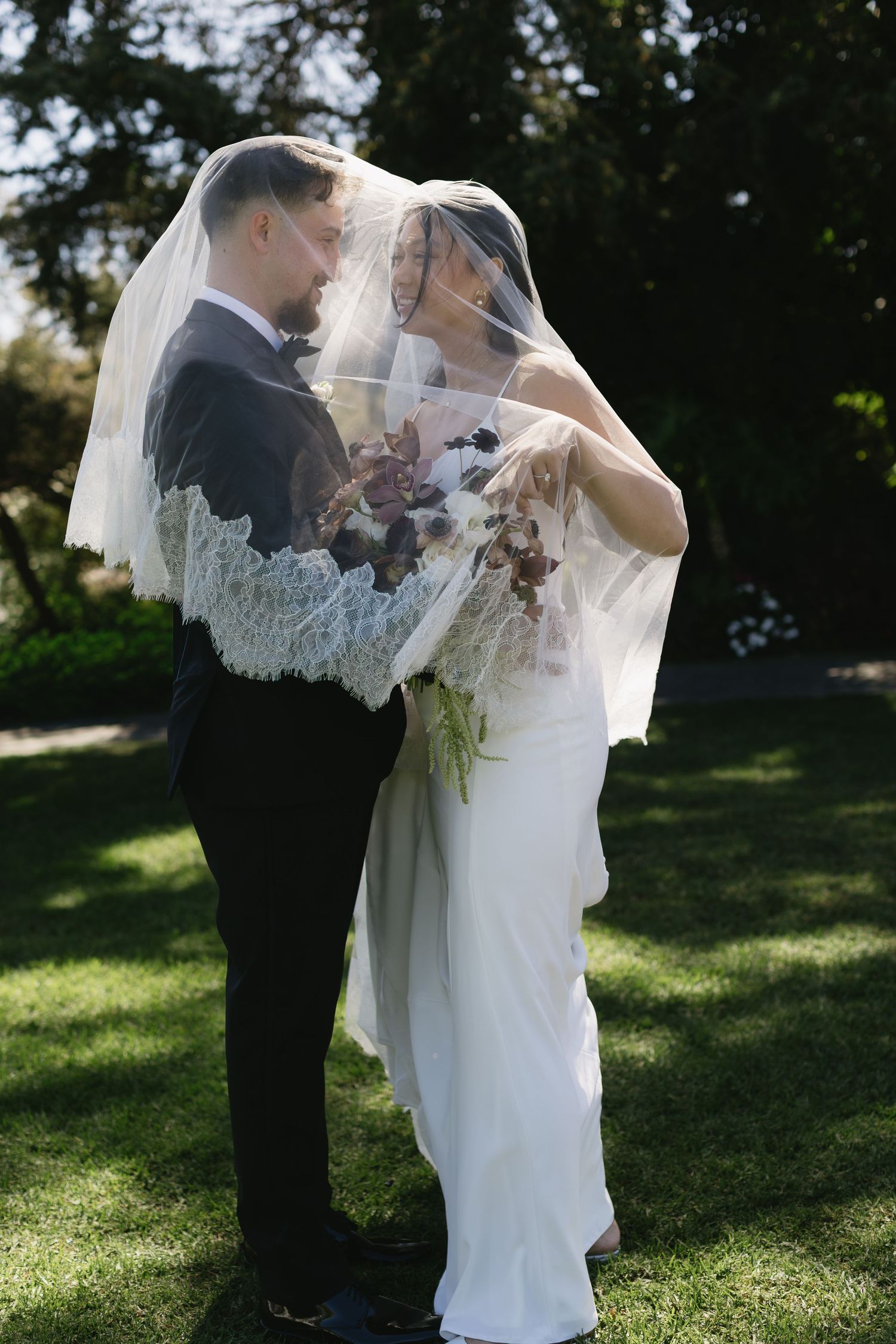 Wedding couple standing together on grass with bride's veil flowing in breeze.