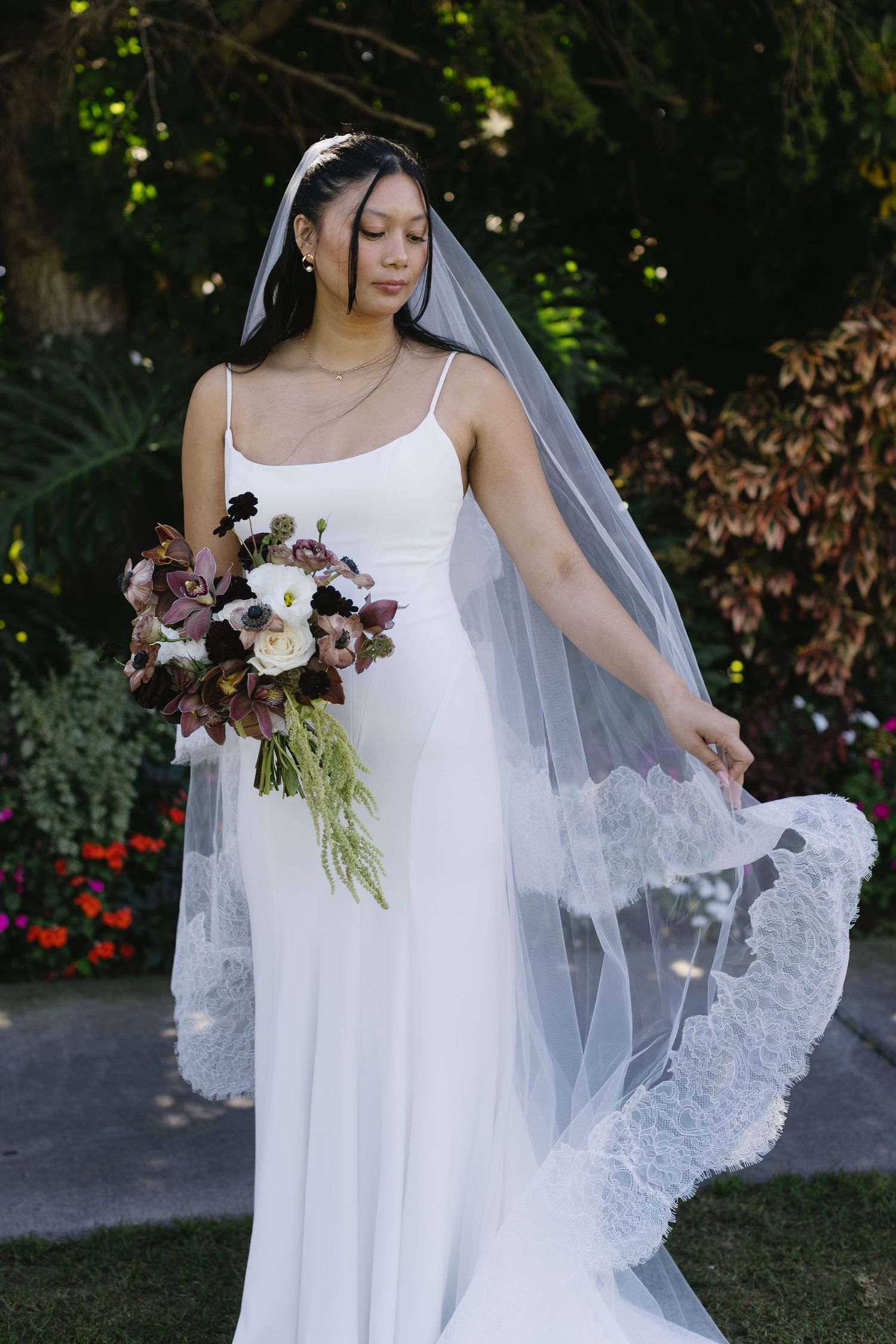 Elegant bride in a white sleeveless gown holding a burgundy and white bouquet with cascading greenery.