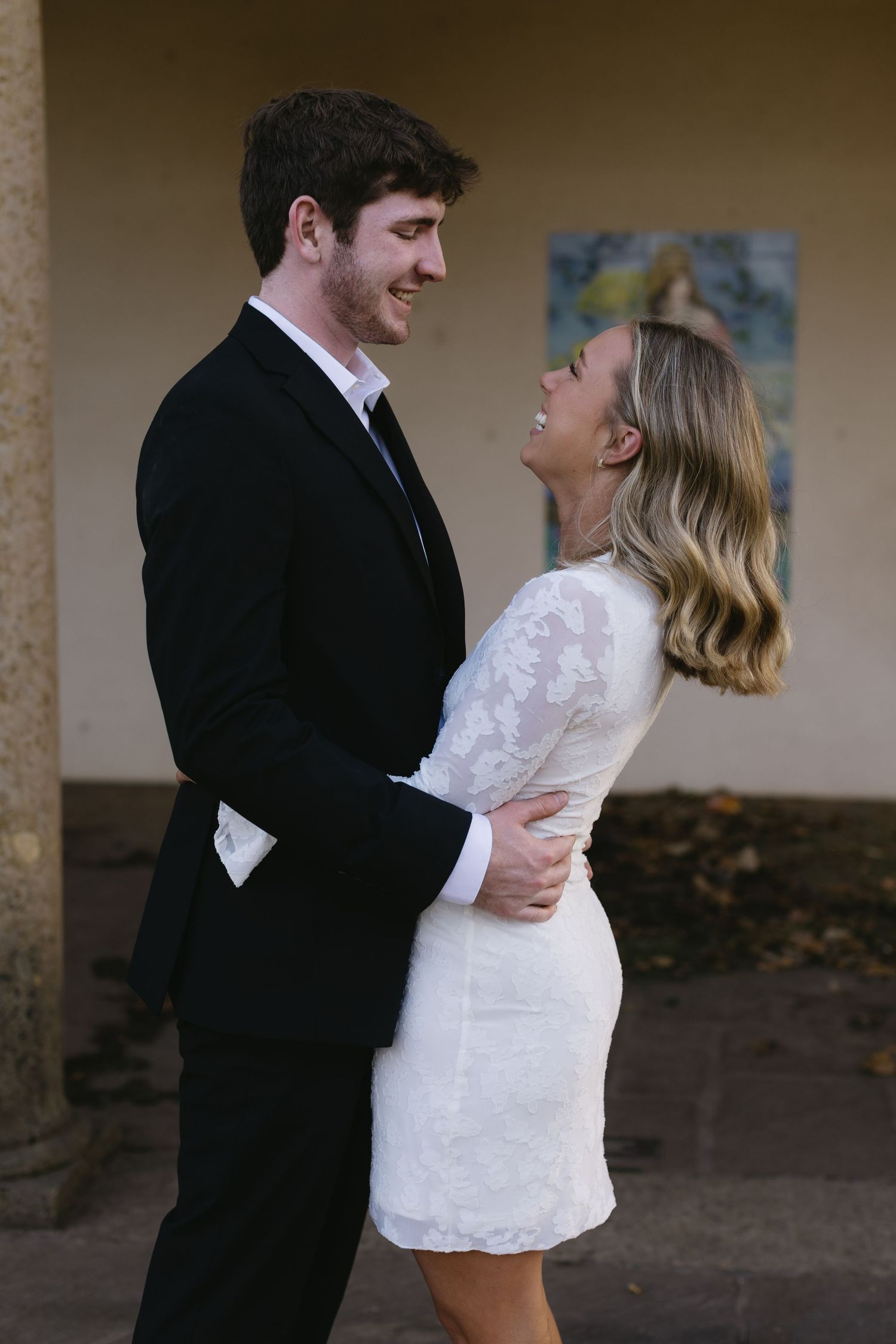 A couple shares an intimate moment, with the bride wearing a short lace dress and groom in formal black suit.