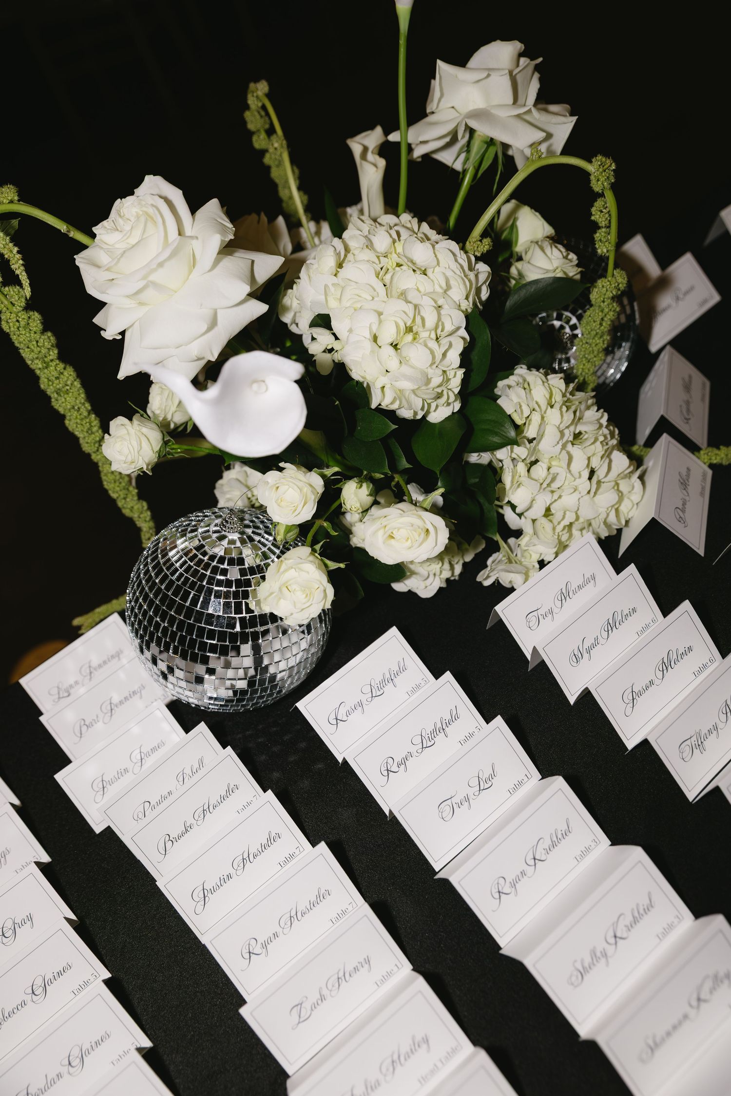 White place cards and floral arrangements with a disco ball on dark surface at wedding reception.