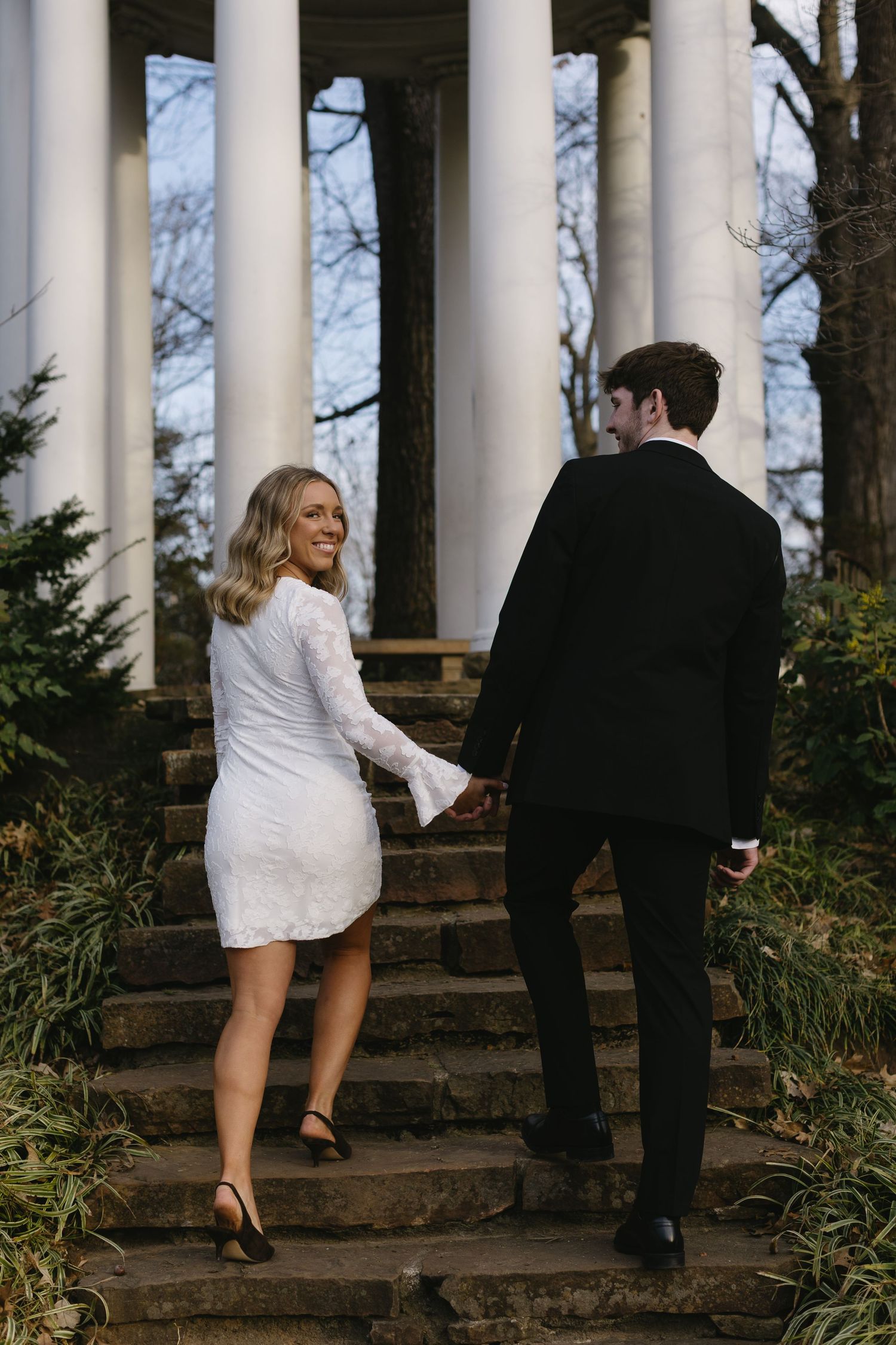 Couple walking up stone steps between white columns in formal attire.