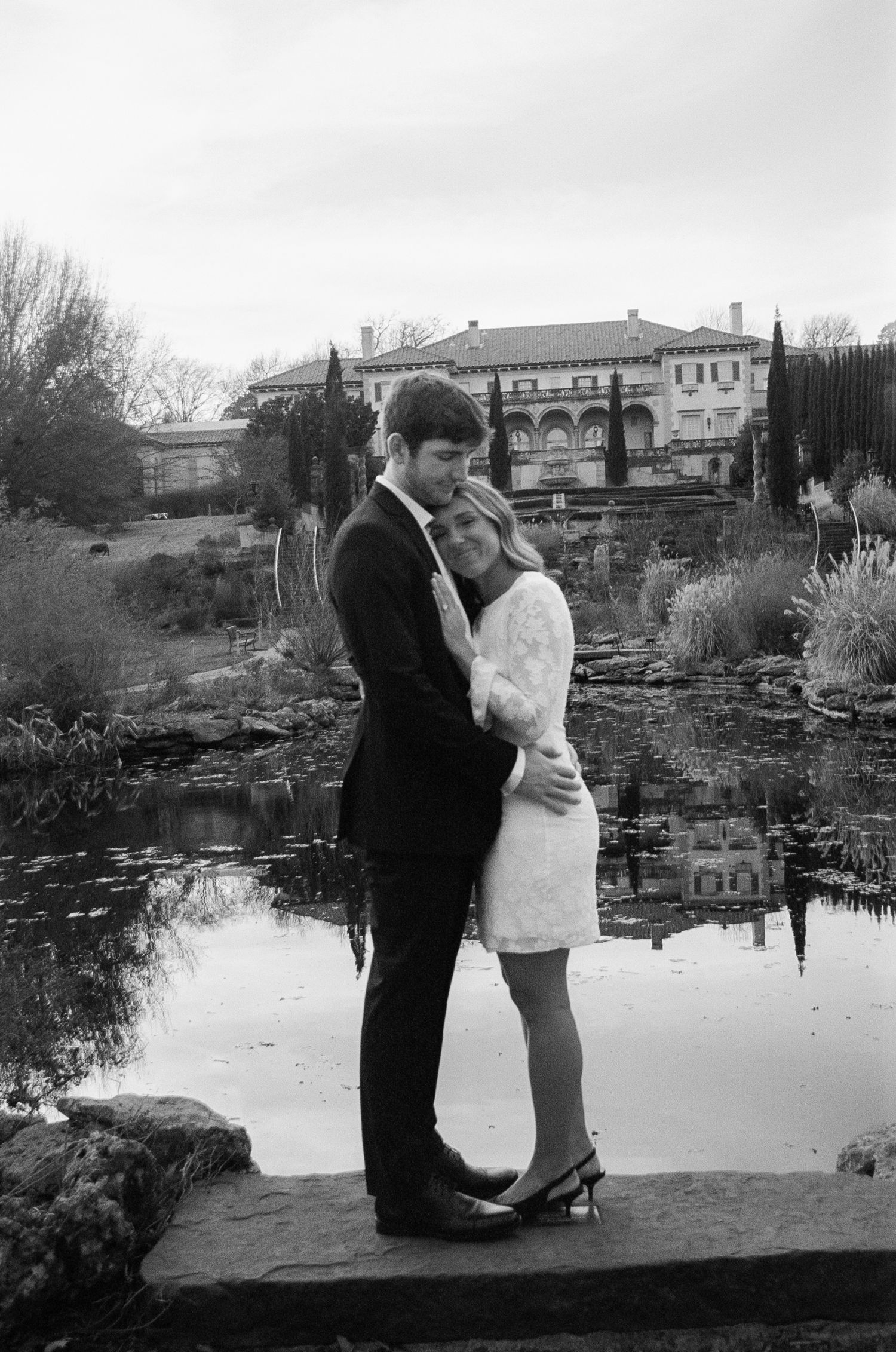 A couple embracing by a reflective pond with historic buildings in the background.