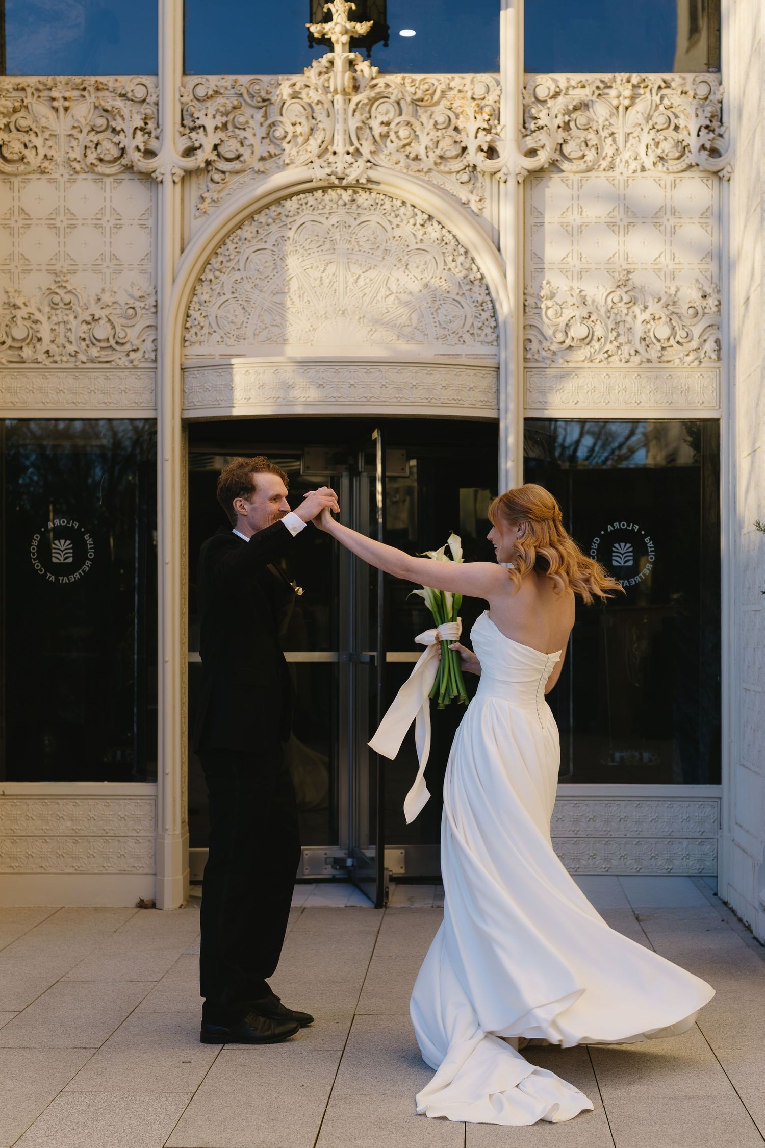 A couple dances playfully outside an ornate building with decorative stone architecture.