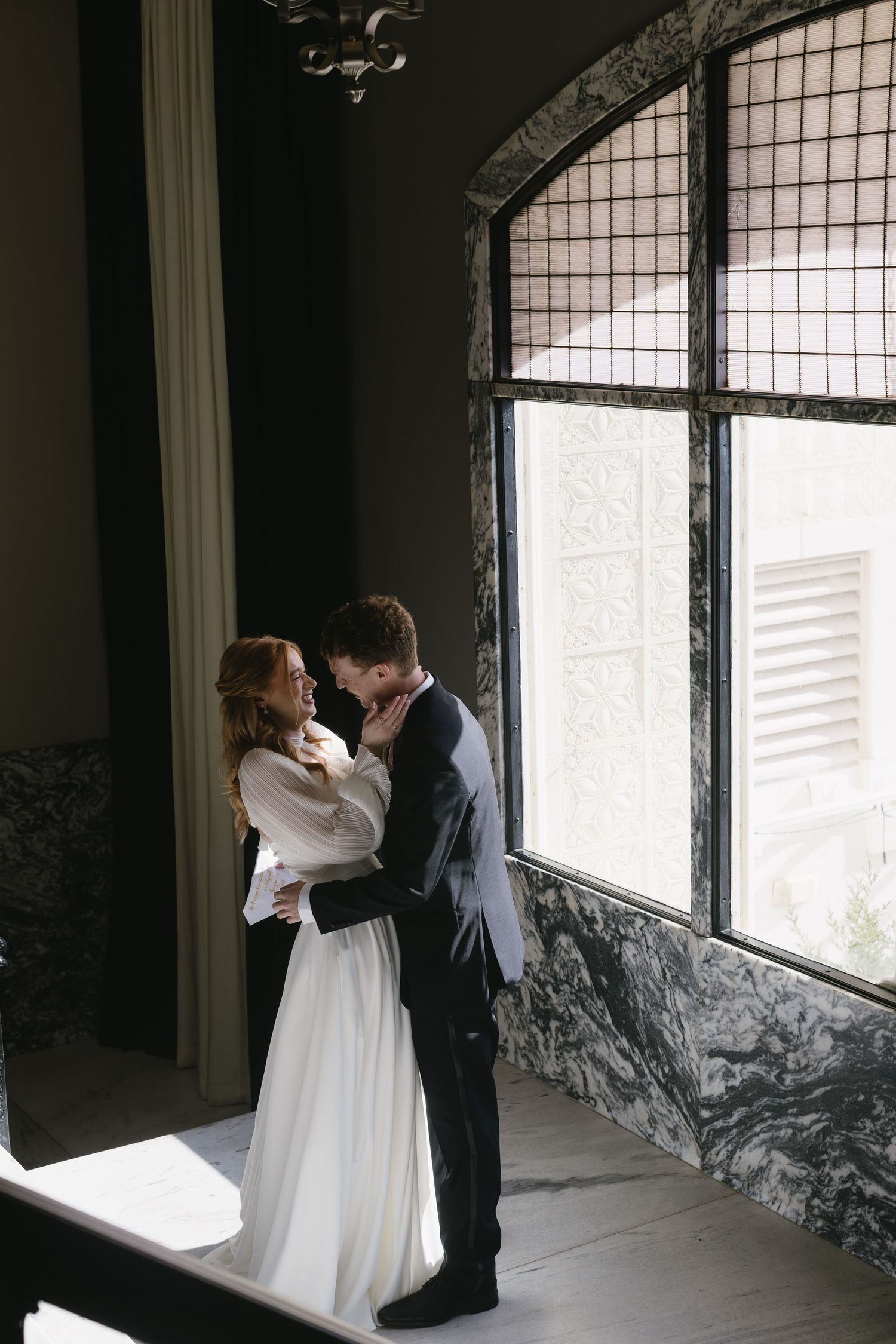 A romantic wedding couple embracing in a windowed hallway with marble walls.