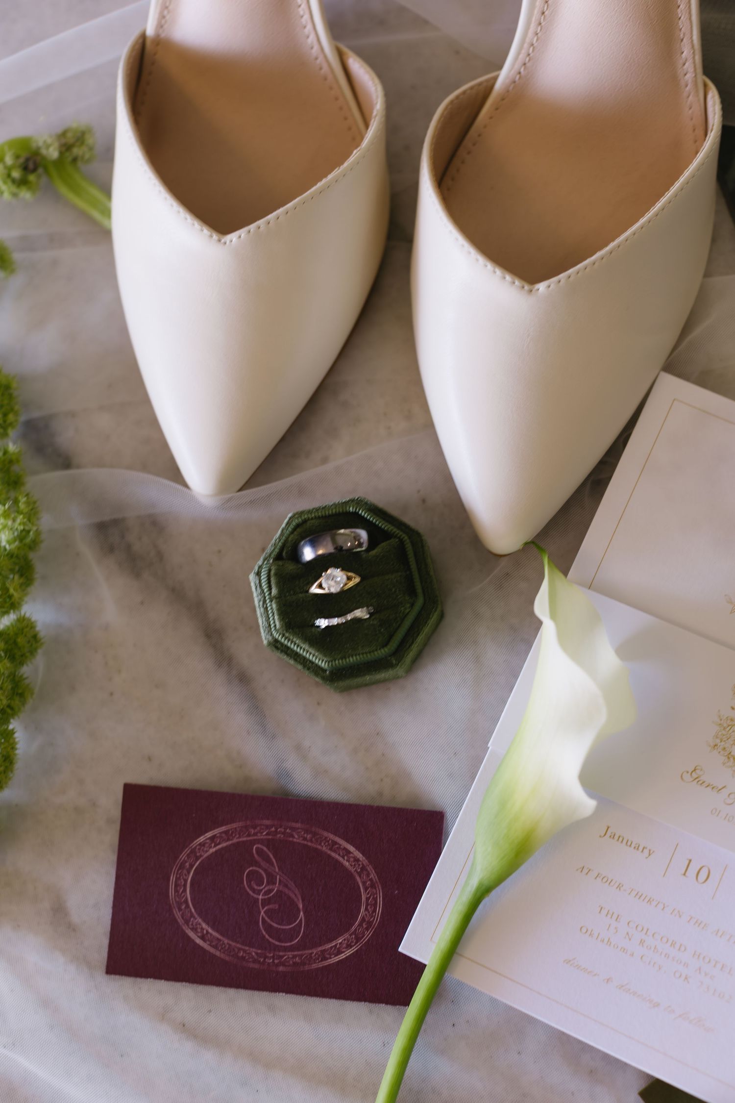 White bridal shoes and wedding accessories arranged on marble surface with purple stationery and green floral accents.