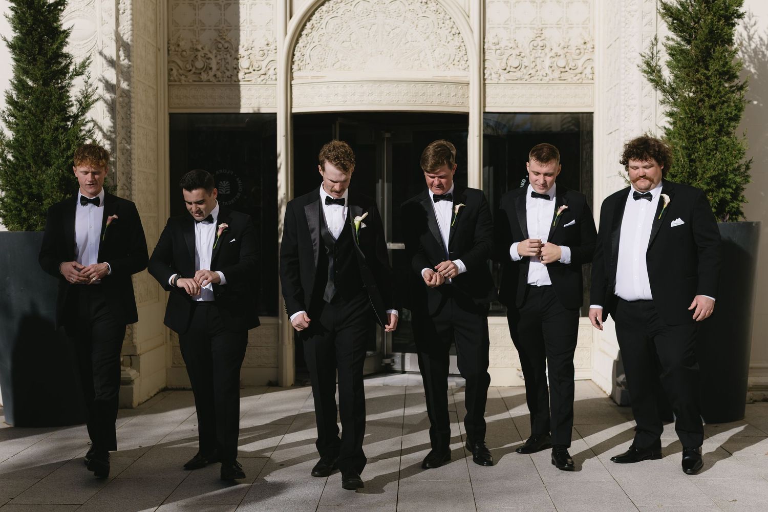 Groomsmen in black tuxedos line up for formal wedding photos outdoors.
