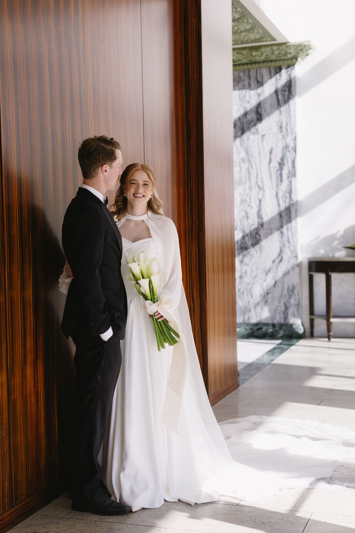 Bride in flowing white gown and groom in tuxedo pose against wooden paneled walls in natural light.