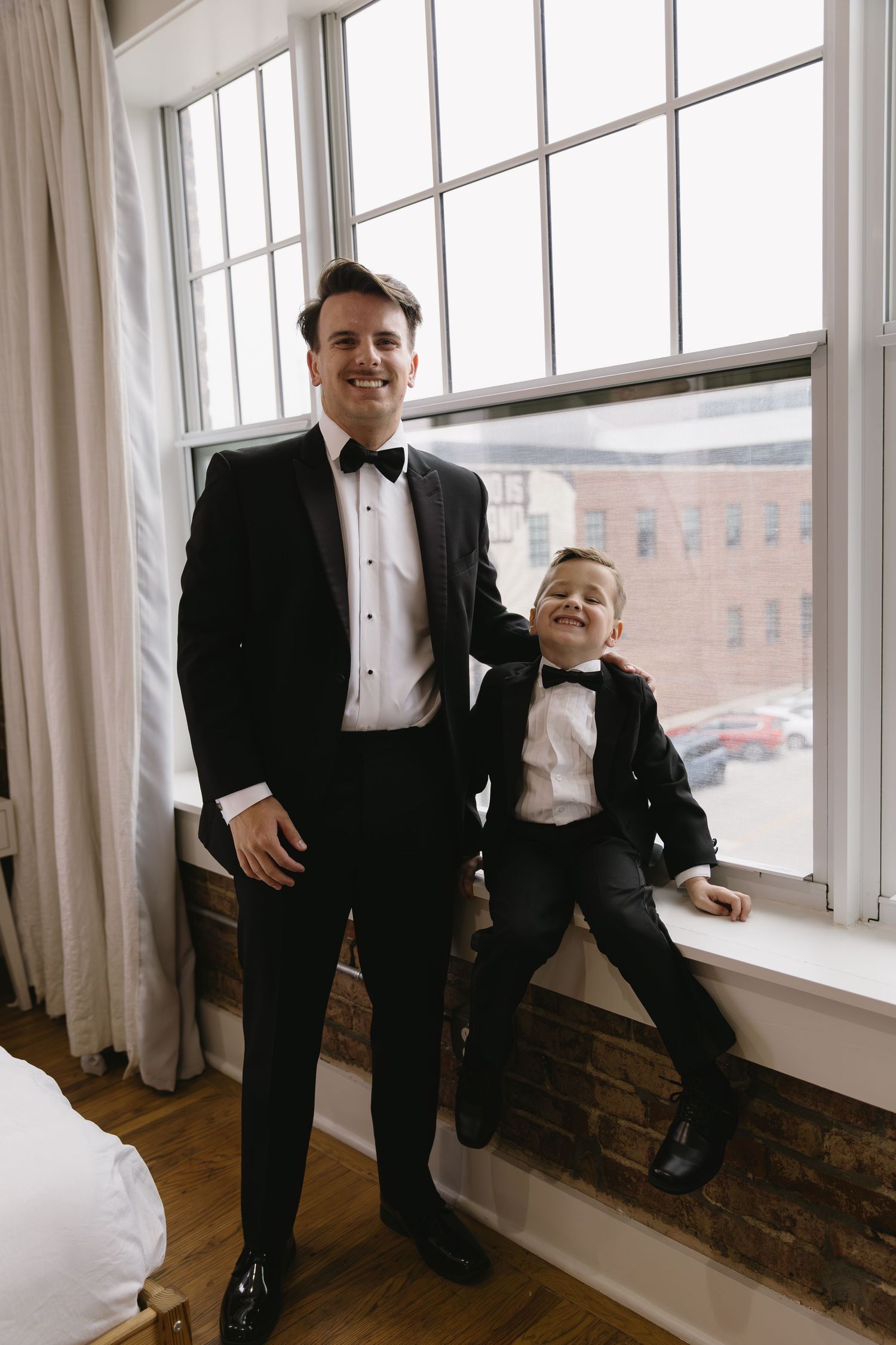 Groomsmen in black tuxedos stand by bright windows in an industrial loft space, creating candid moments.
