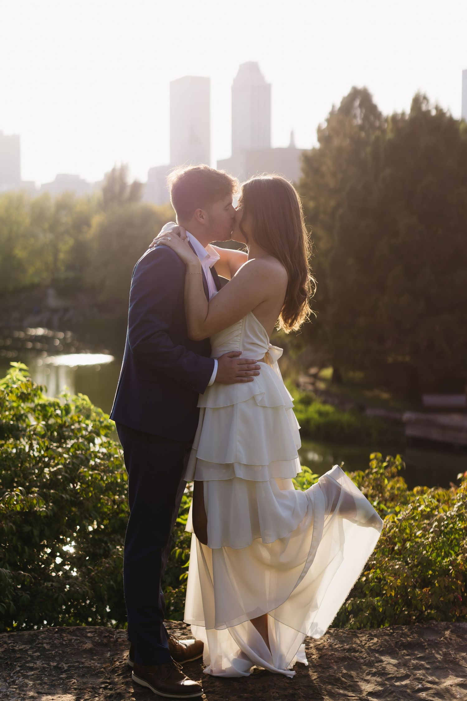 A couple embraces in a sunlit garden with city skyline visible in the background.