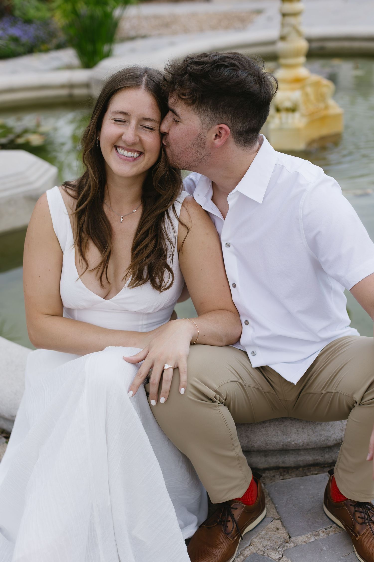 A couple shares an intimate moment while sitting by a decorative fountain in a garden.