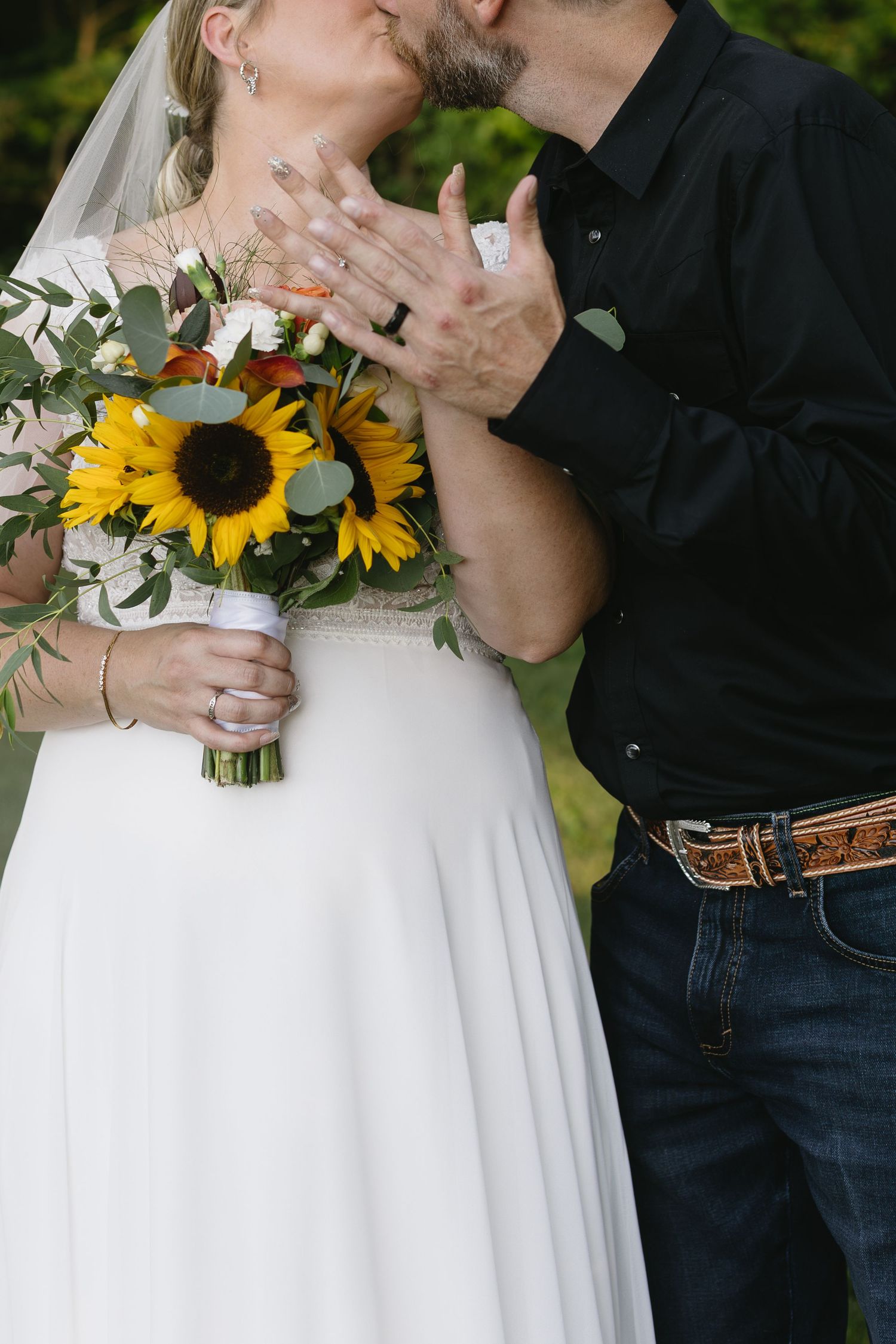 A wedding couple holds hands while showcasing a beautiful sunflower bouquet against a natural outdoor backdrop.
