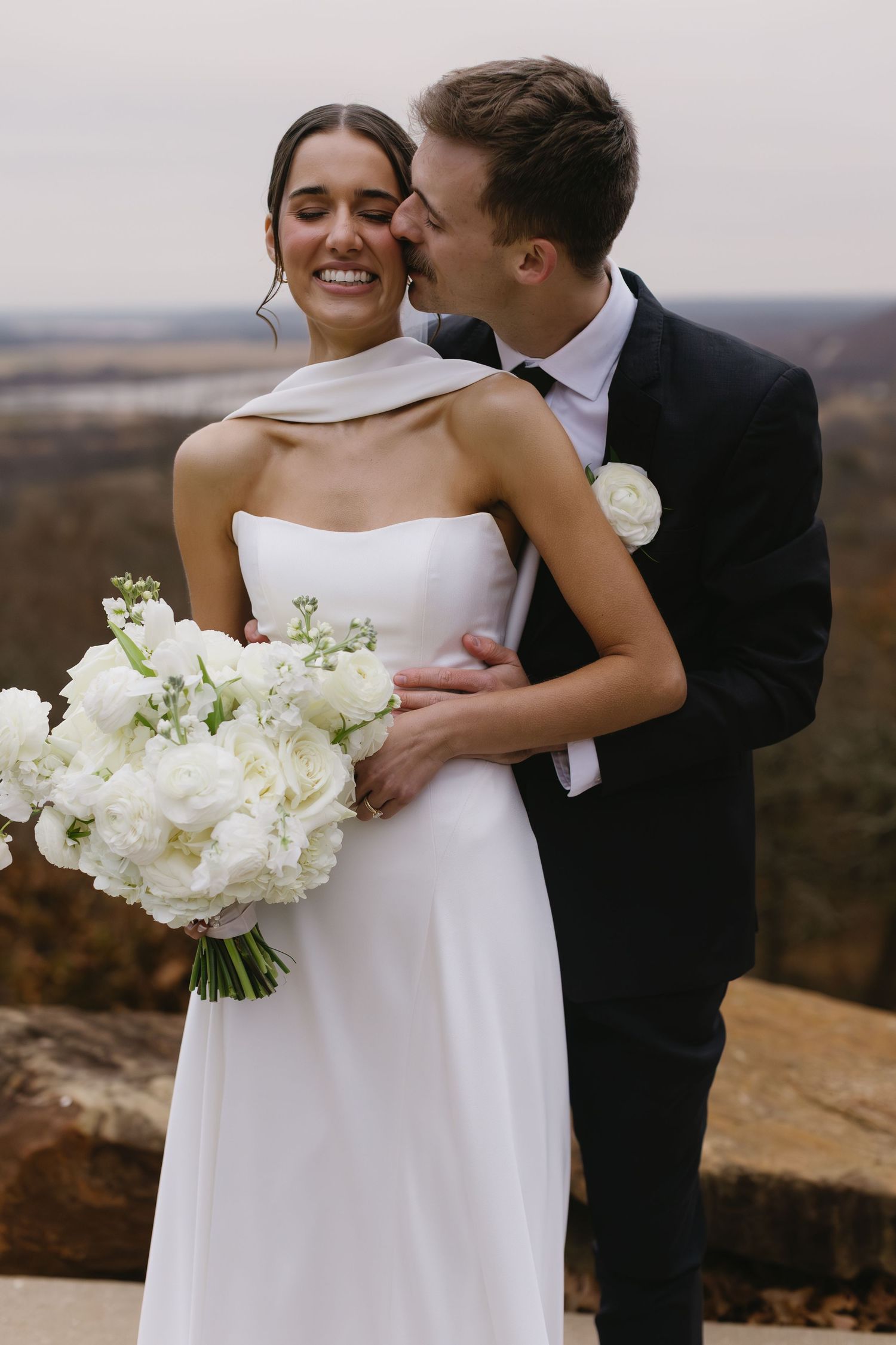 Romantic wedding couple embraces on a scenic cliff overlook with a white floral bouquet.