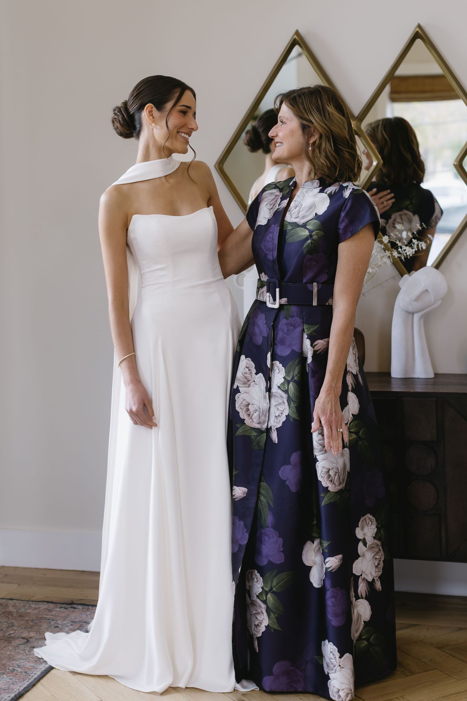 Two women stand together, one in a white wedding gown and another in a purple floral dress.