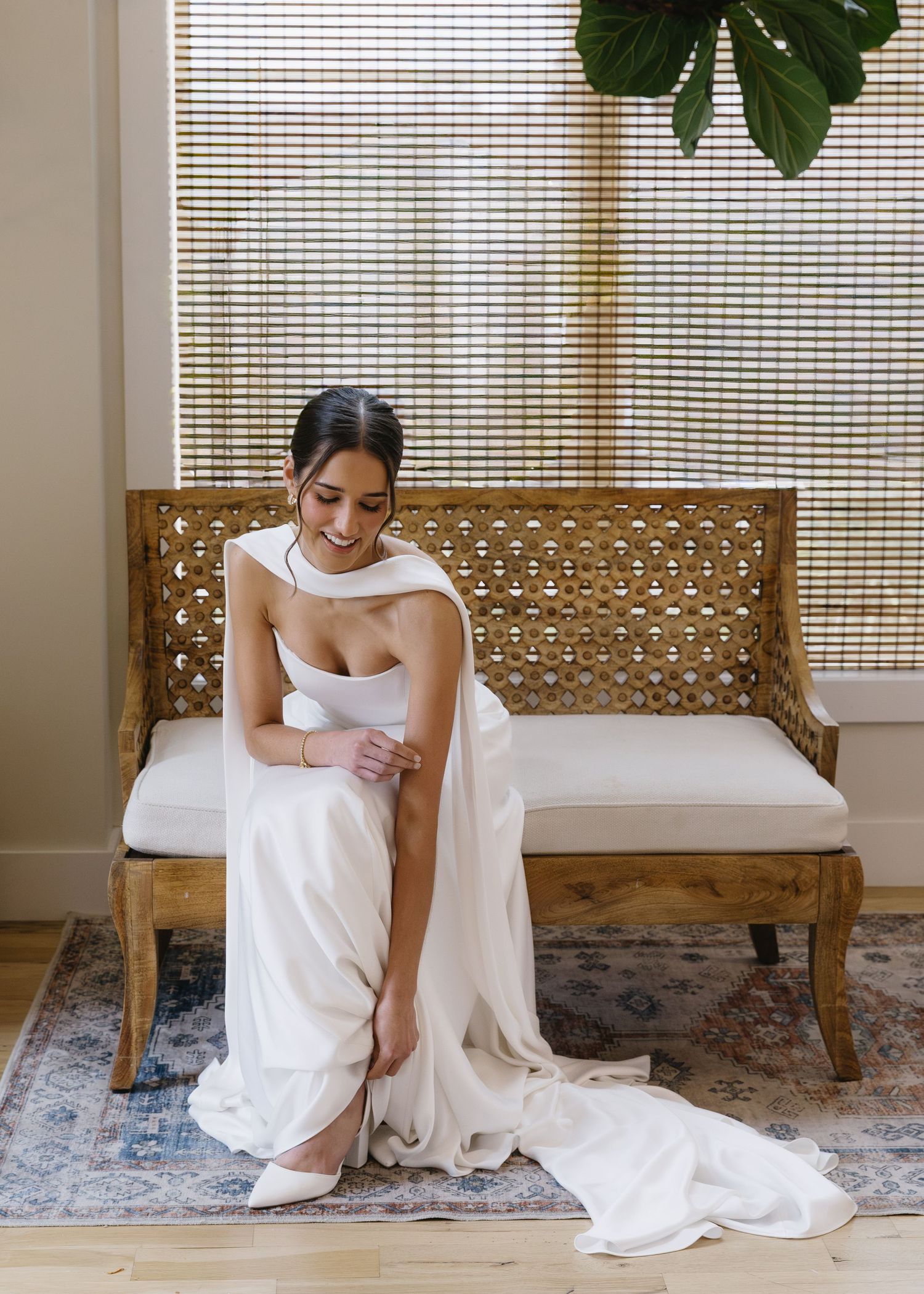 Bride in white dress sitting on woven bench in sunlit room with window blinds.