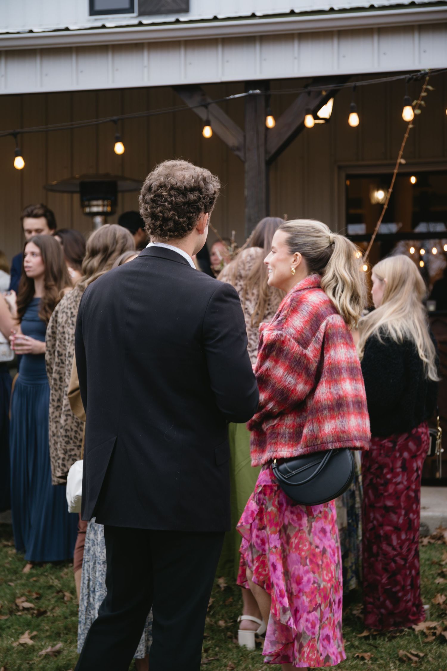 Wedding guests in formal attire socialize outside a venue with string lights.