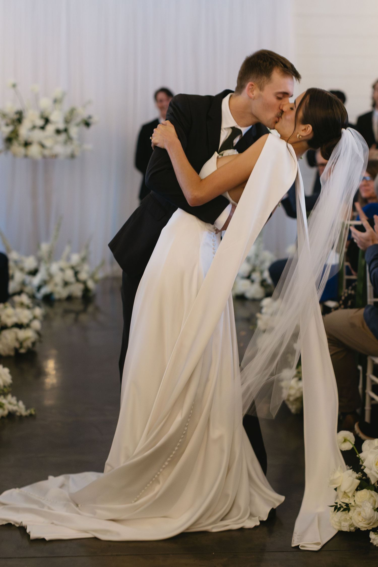Romantic wedding scene with couple in white dress and black suit sharing a dip kiss.