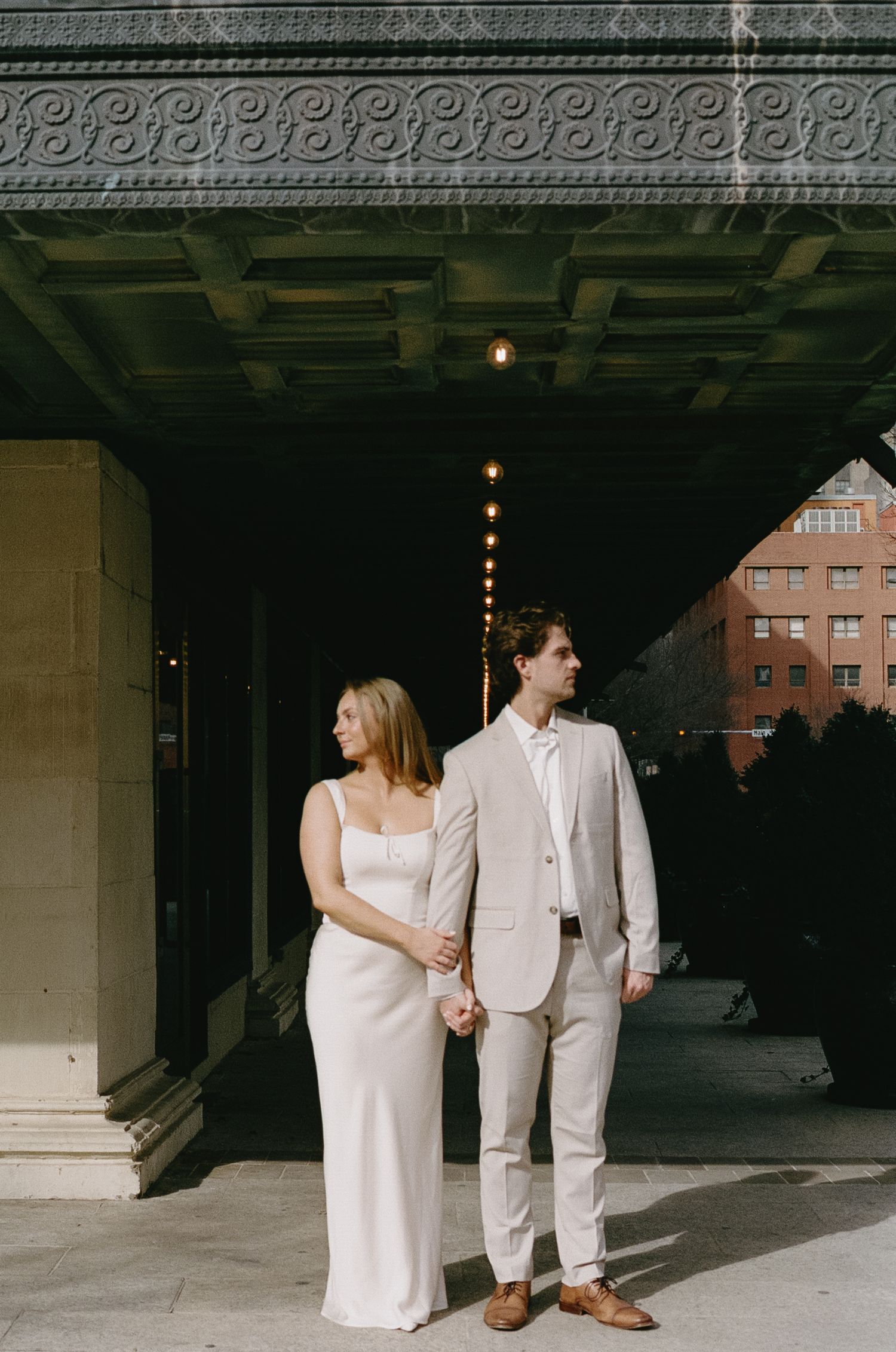 Elegant couple in white attire posing under architectural details of urban building.