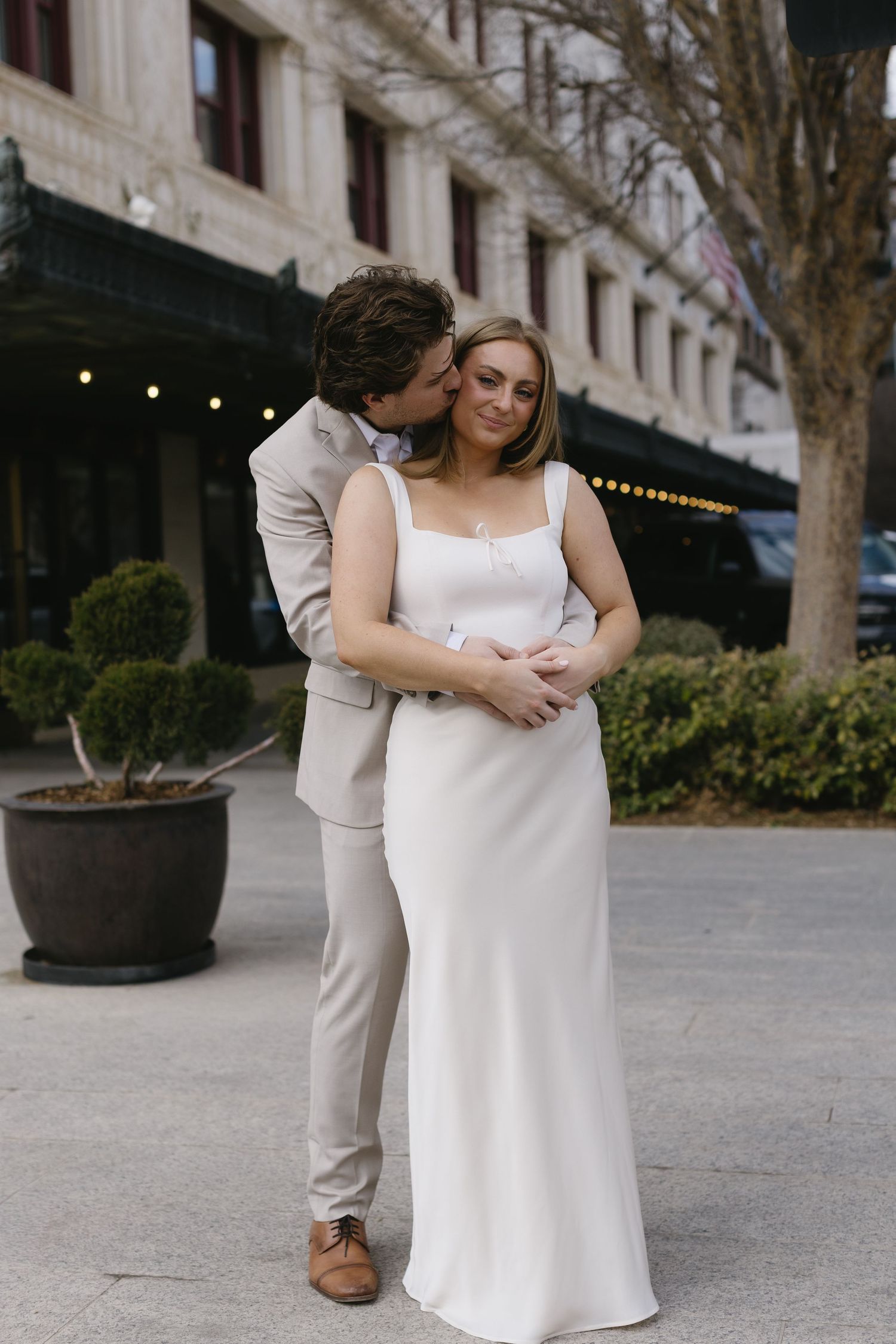 A couple in coordinated white and beige outfits embrace tenderly on an outdoor plaza.