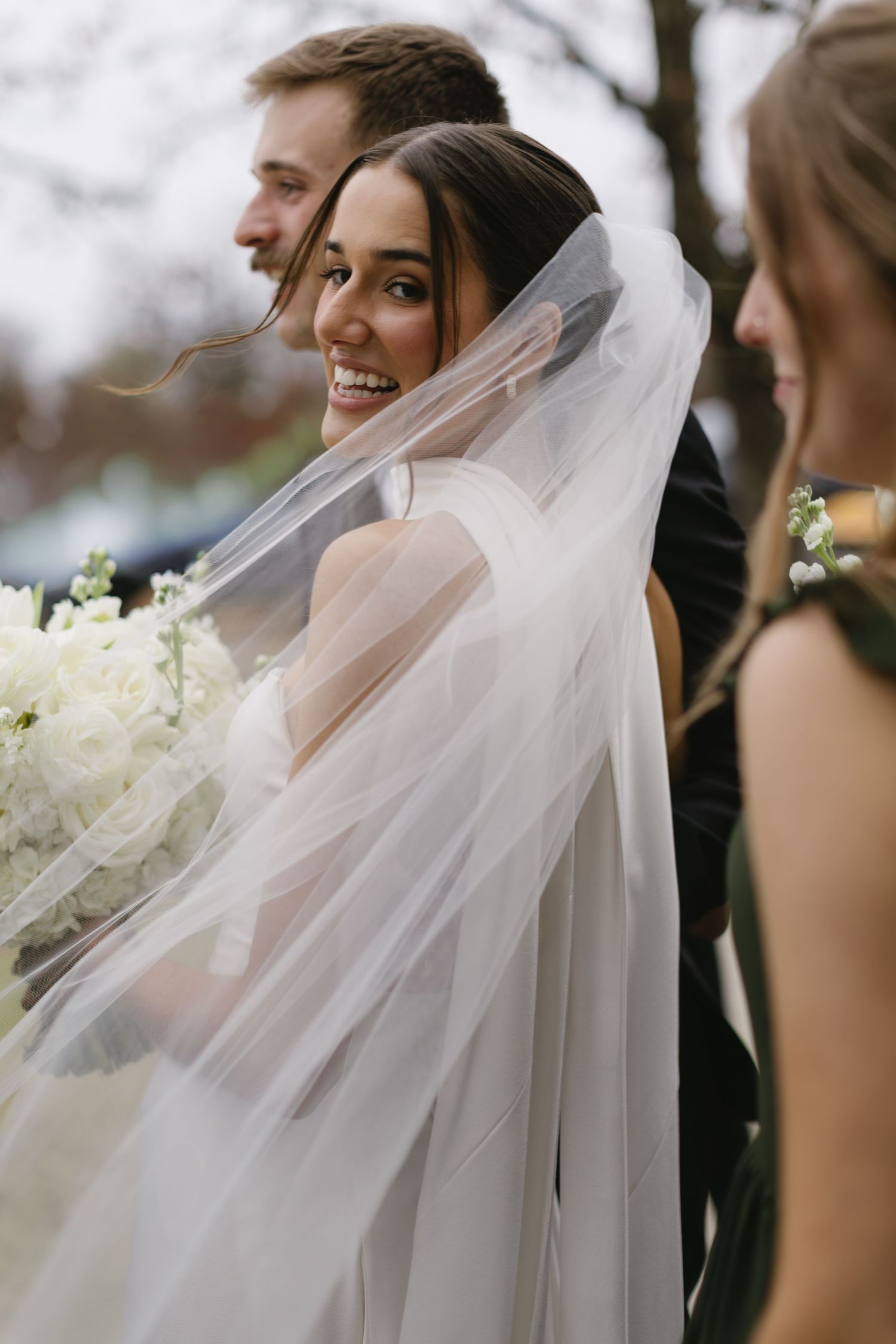 A bride with a flowing veil smiles during outdoor wedding photos.