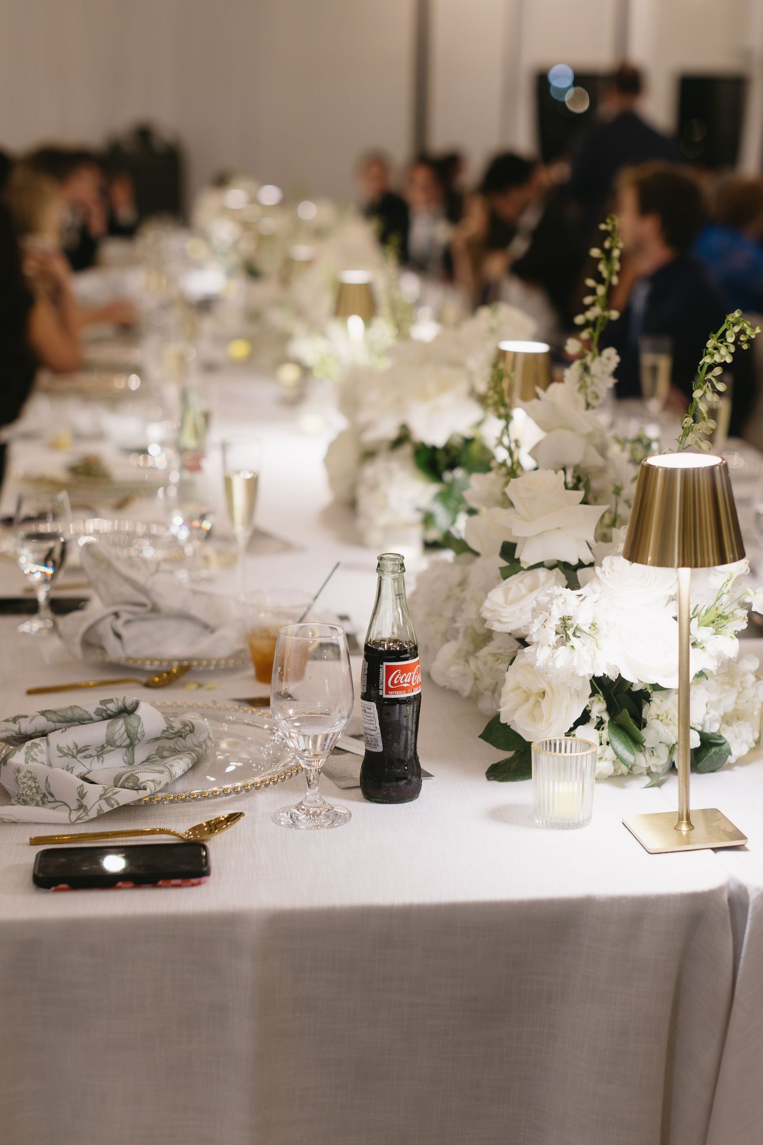 Wedding reception table decorated with white linens, floral centerpieces, and miniature gold lamps.