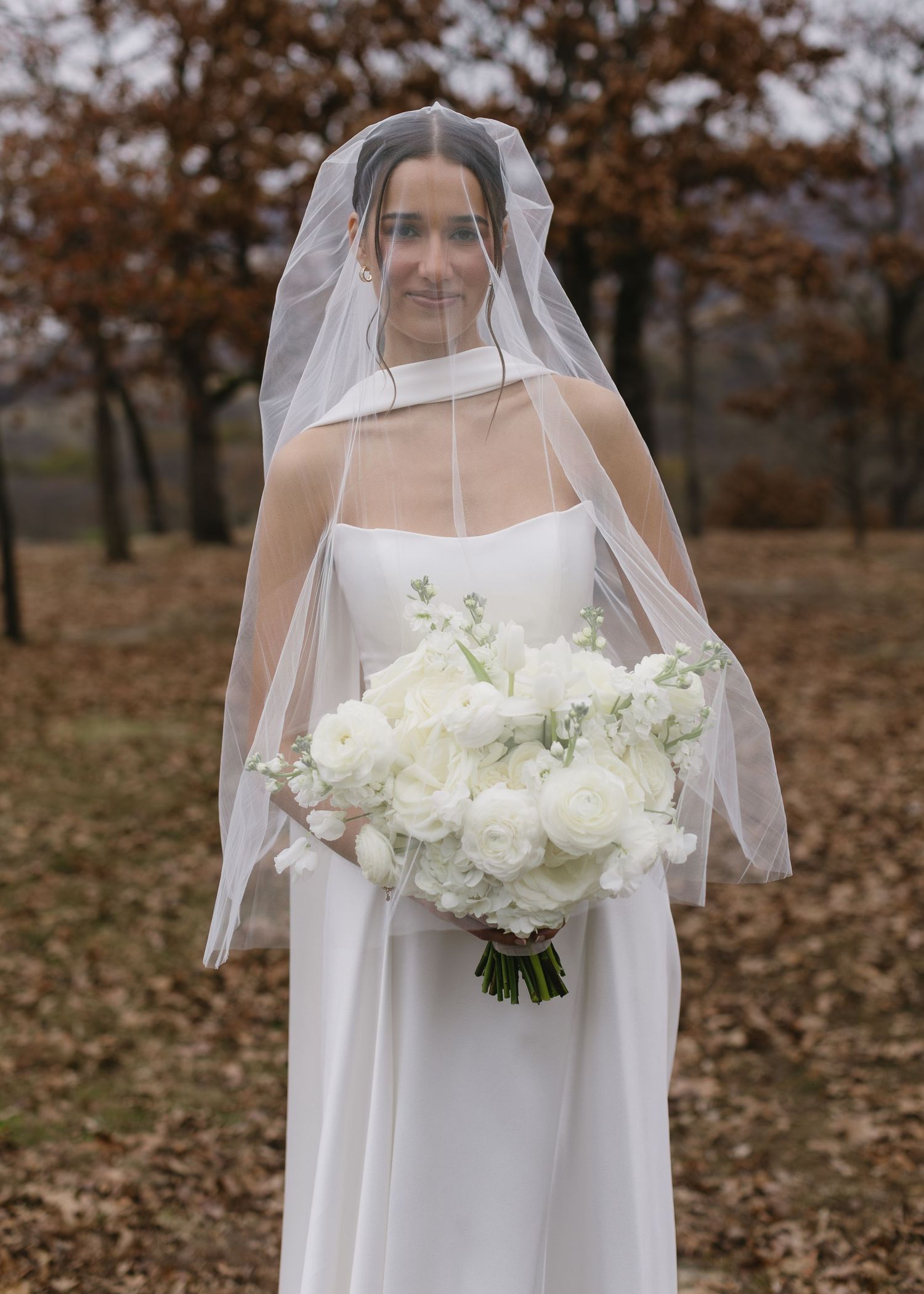 A bride in a classic white gown holds a white bouquet while wearing a traditional veil outdoors.
