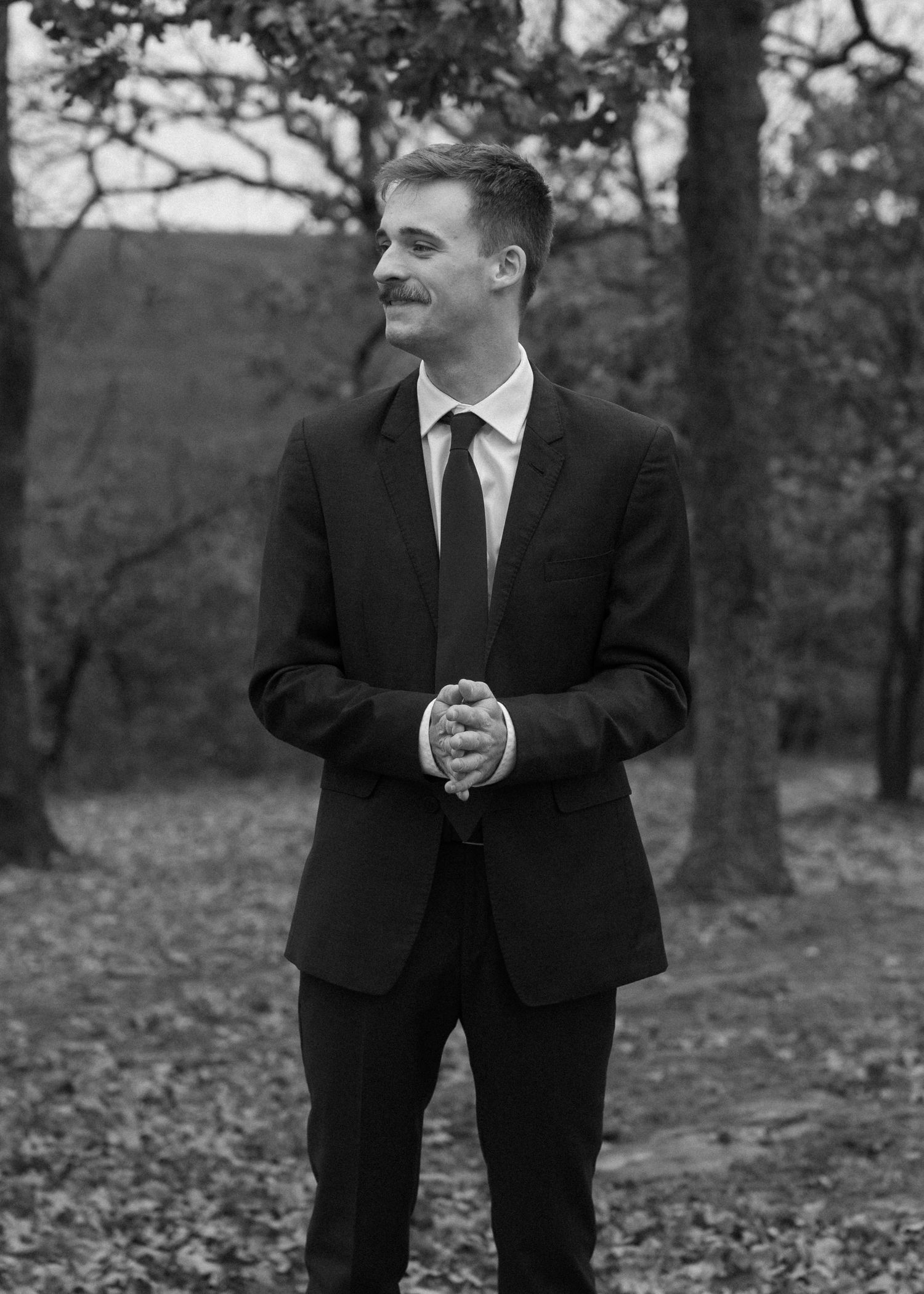 Black and white photo of a groom in a classic suit posing in an autumn forest setting.
