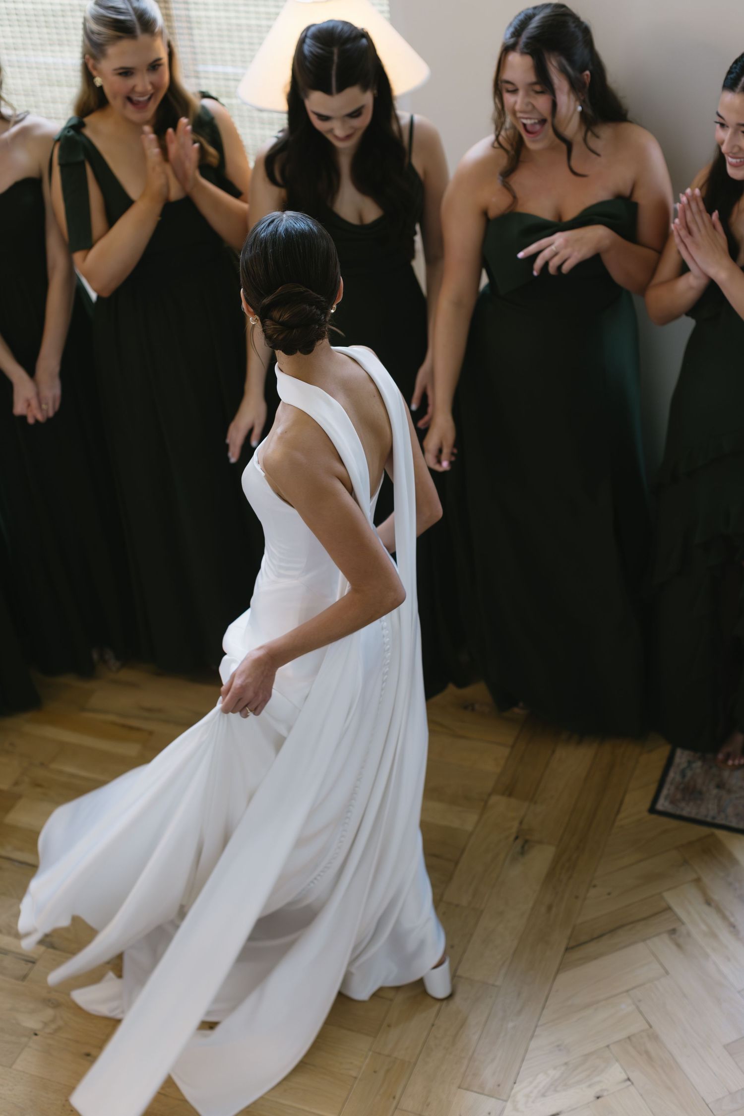 A bride in white twirls her dress while surrounded by bridesmaids in black gowns.