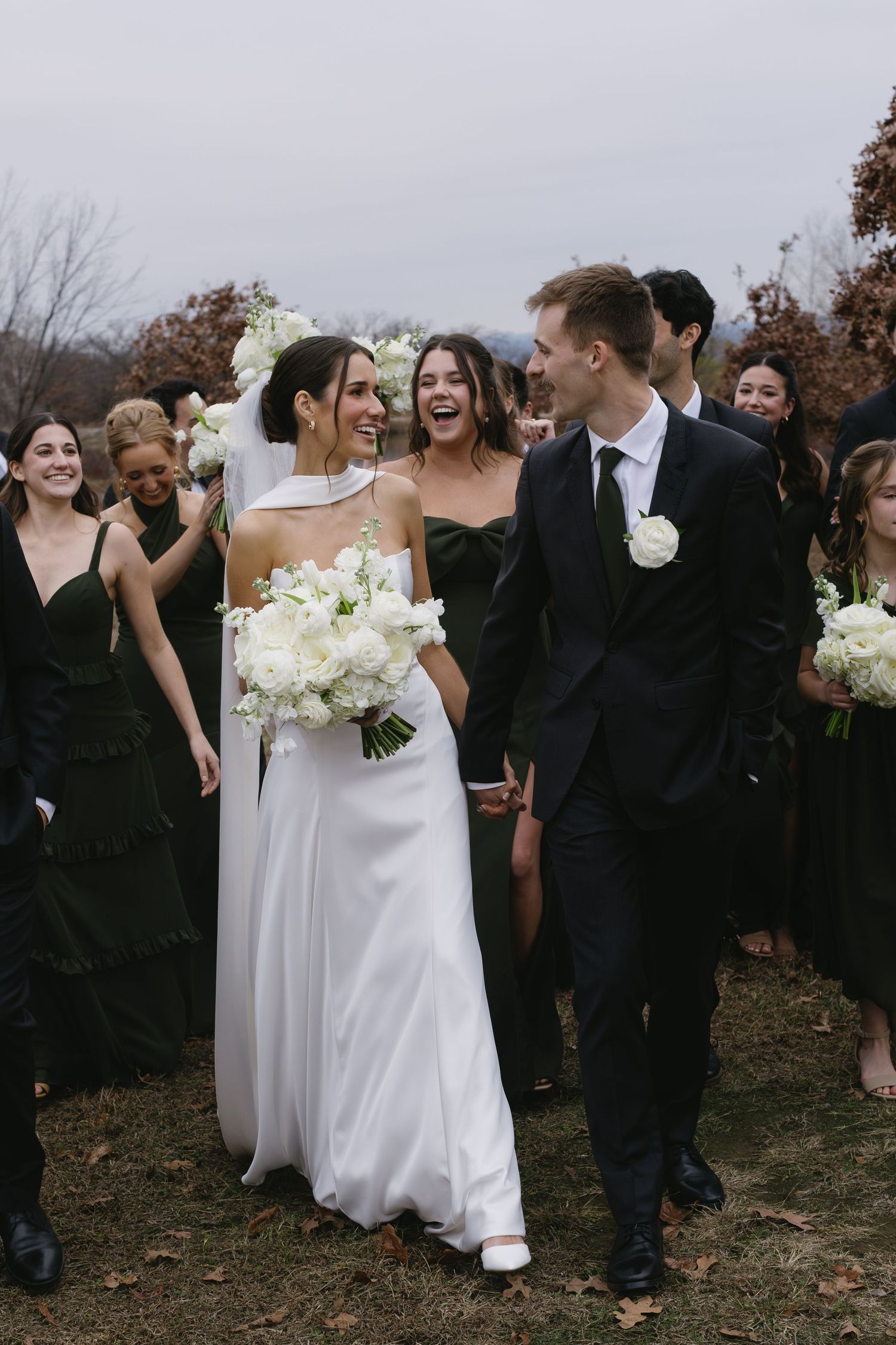 Wedding party walking together outdoors with bridesmaids in dark dresses and white bouquets.