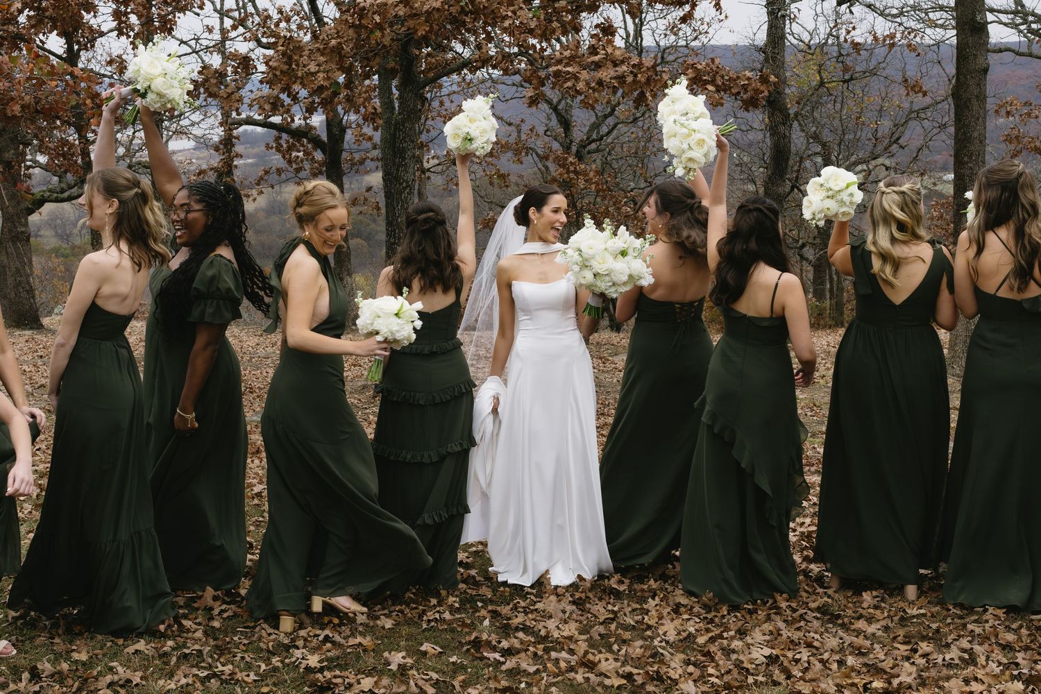 Large wedding party in black attire poses for group photos in an autumn forest setting.
