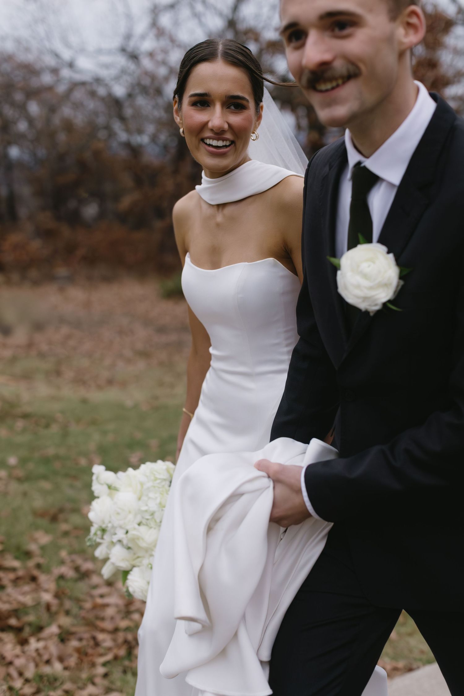 Elegant bride in white dress and groom in black tuxedo embrace during outdoor autumn wedding photoshoot.