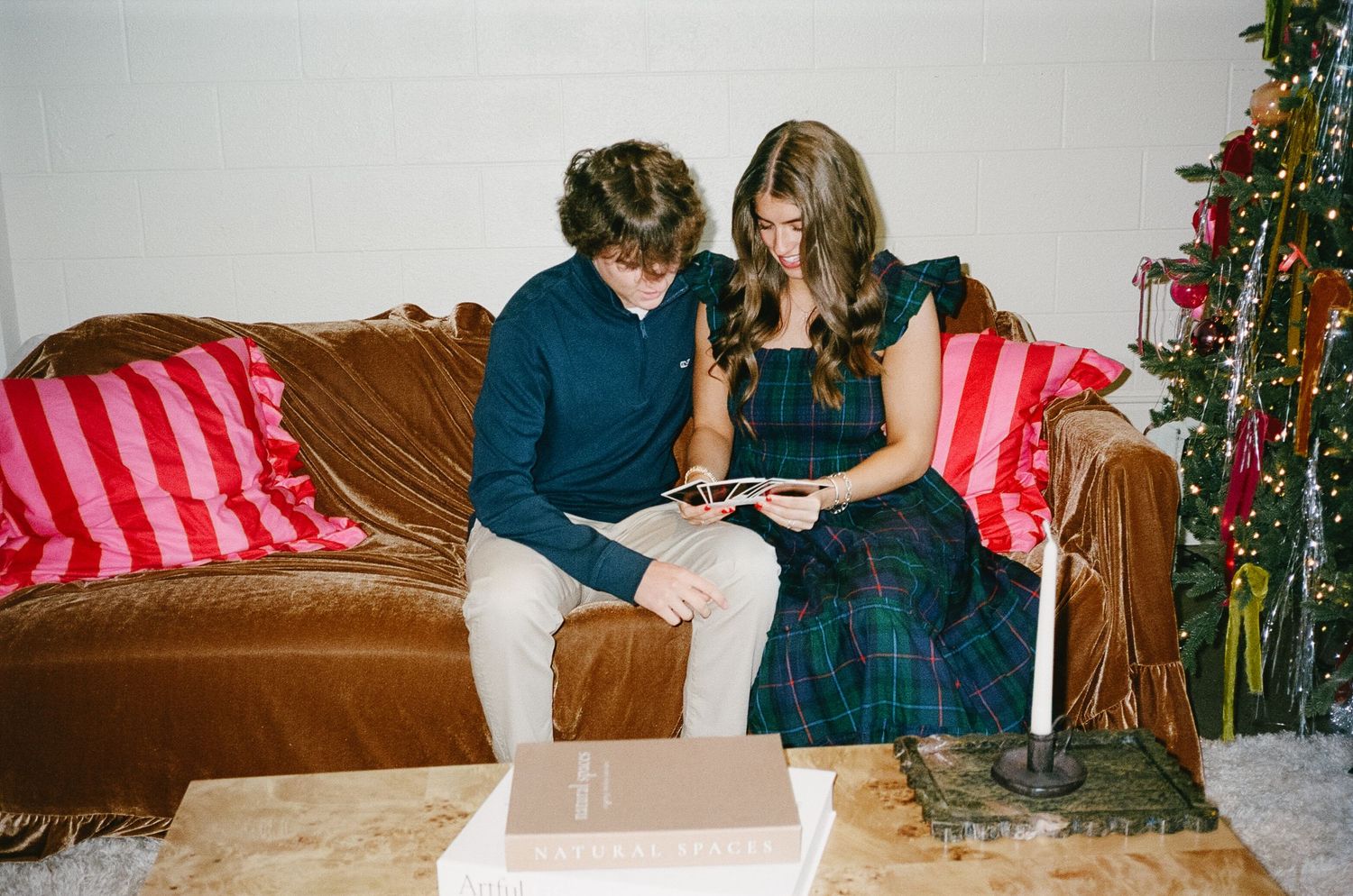 Two people sit together on a brown leather couch looking at a book near a decorated Christmas tree.