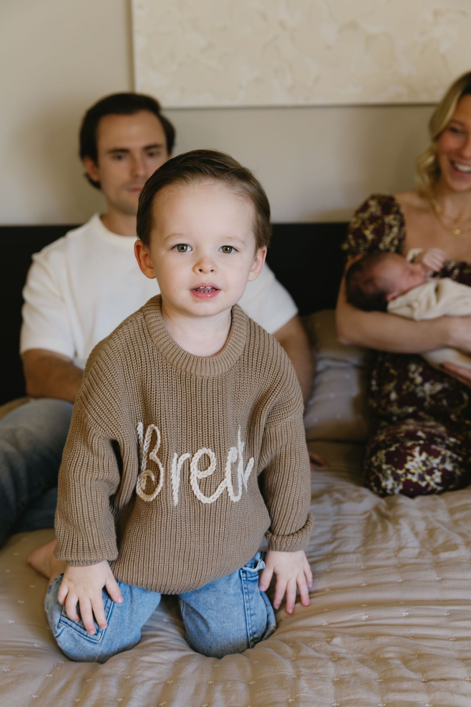Small child in brown sweater and jeans sitting on bed with family members in background.